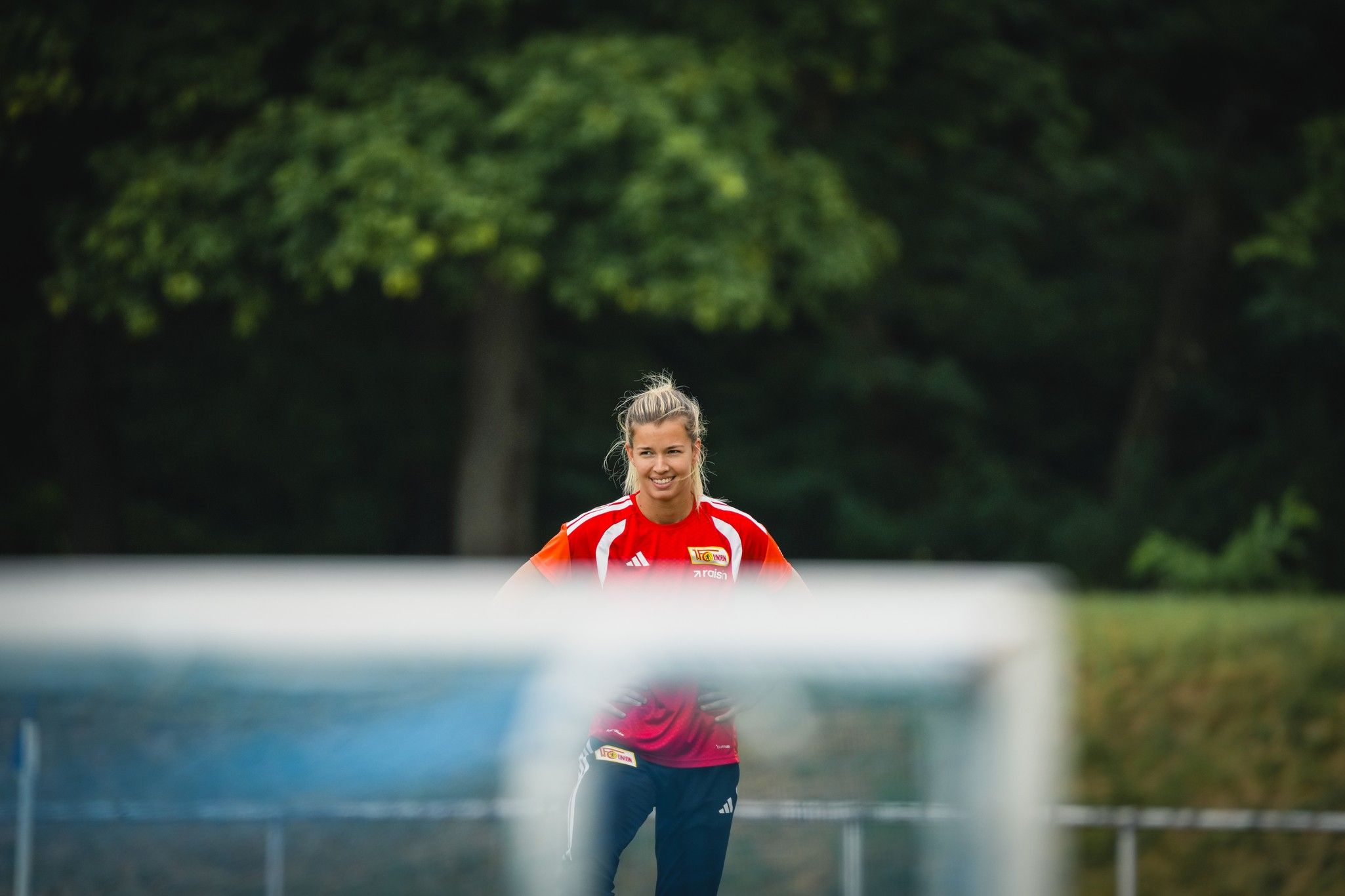 Eine lächelnde Frau in rotem Trikot steht auf einem Fußballplatz, im Hintergrund Bäume und ein Fußballtor unscharf.
