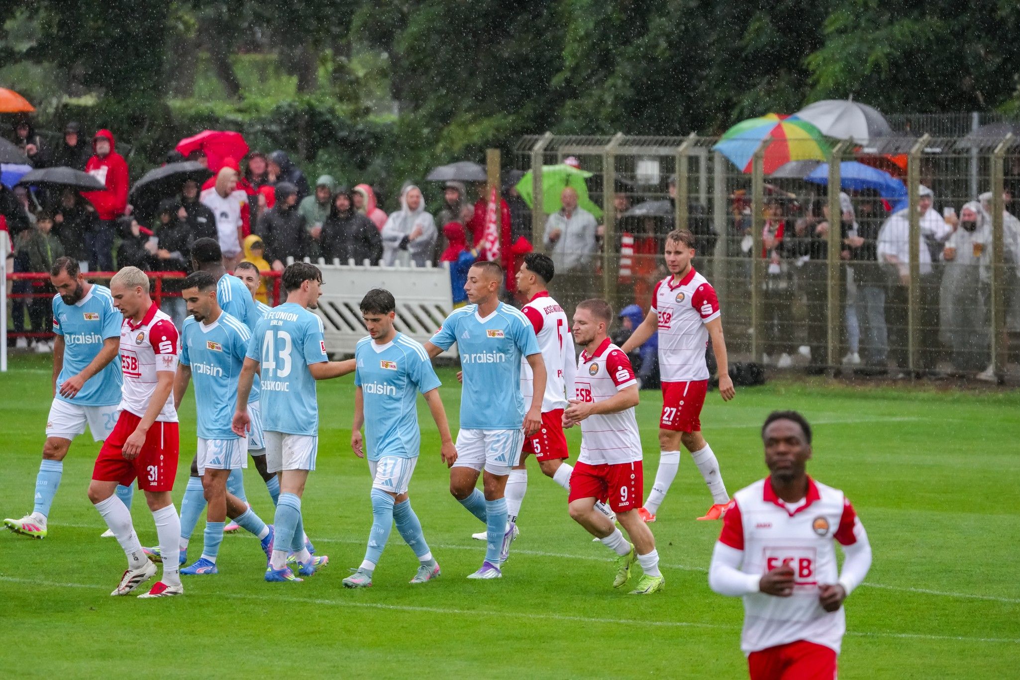 Spieler in blauen und roten Trikots auf dem Platz, während es regnet. Zuschauer mit Regenschirmen im Hintergrund.