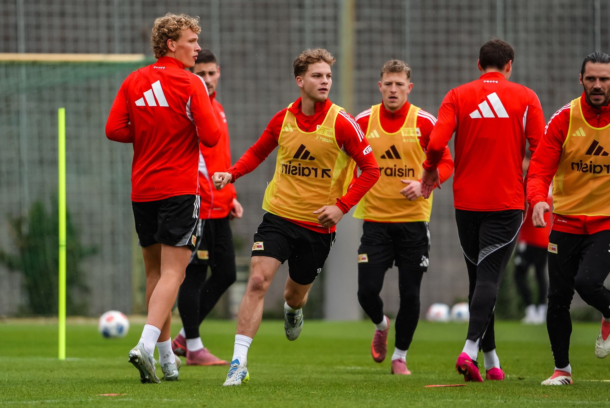 Fünf Fußballspieler in roten Trikots und gelben Trainingswesten trainieren auf einem Rasenplatz.