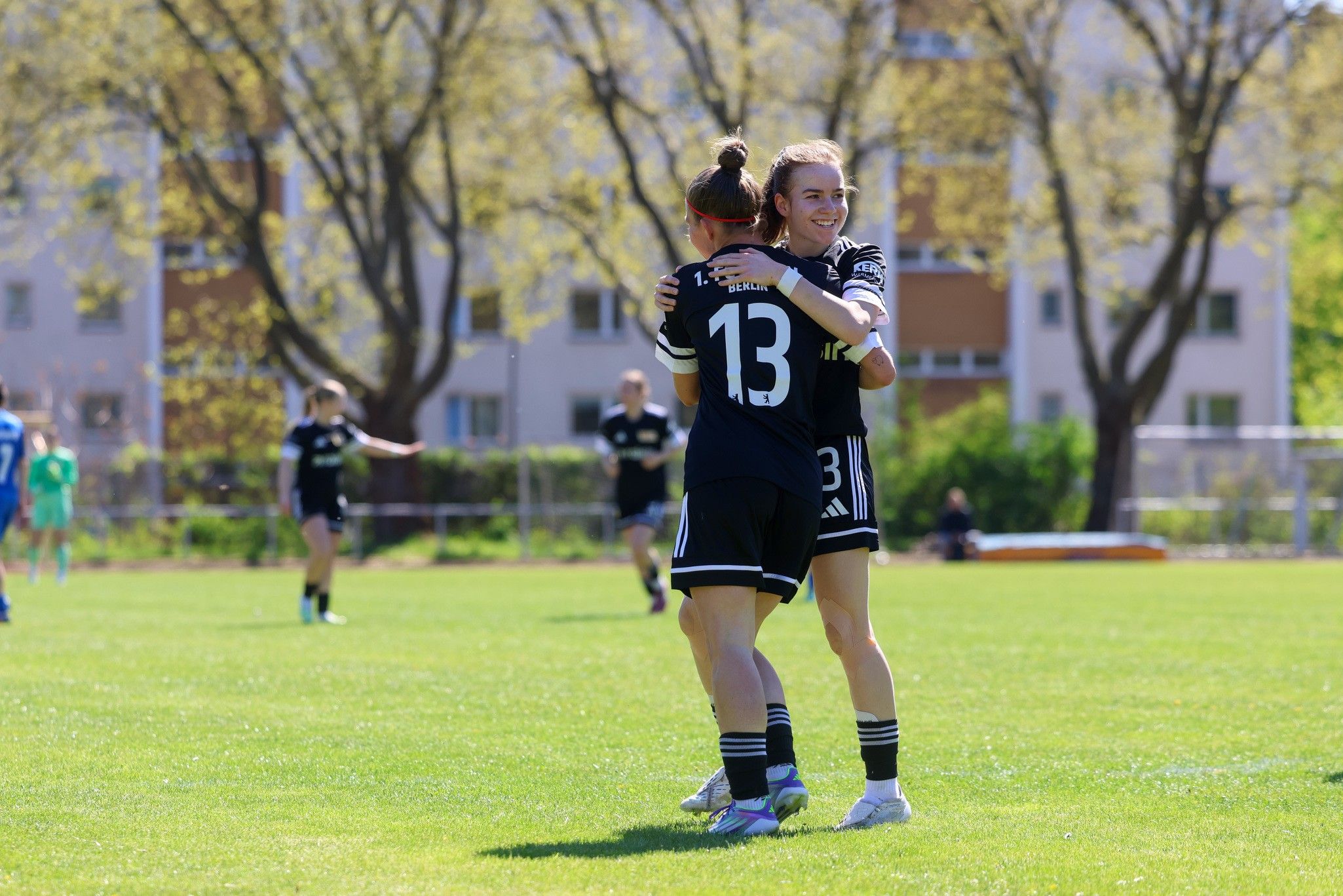 Zwei Fußballspielerinnen in schwarzen Trikots umarmen sich auf einem grünen Feld bei sonnigem Wetter.