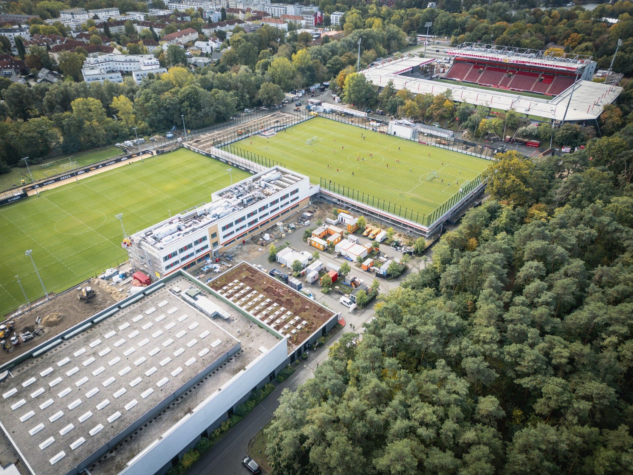 Luftaufnahme eines Sportgeländes mit mehreren Fußballplätzen, umgeben von Bäumen und Gebäuden. Im Hintergrund ein Stadion.