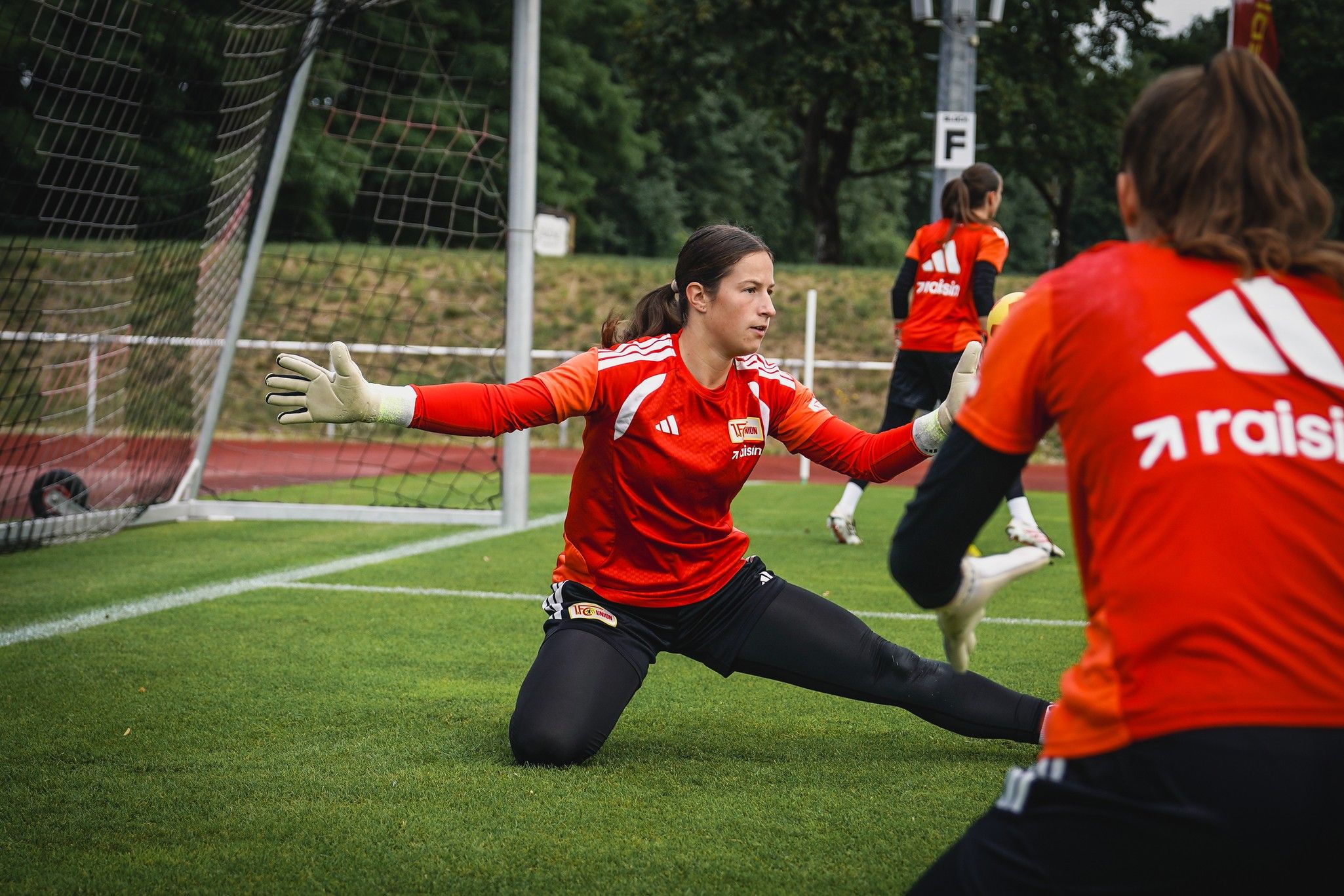 Eine Torhüterin in rotem Trikot übt auf einem Fußballfeld, während eine andere Spielerin im Hintergrund zuschaut.