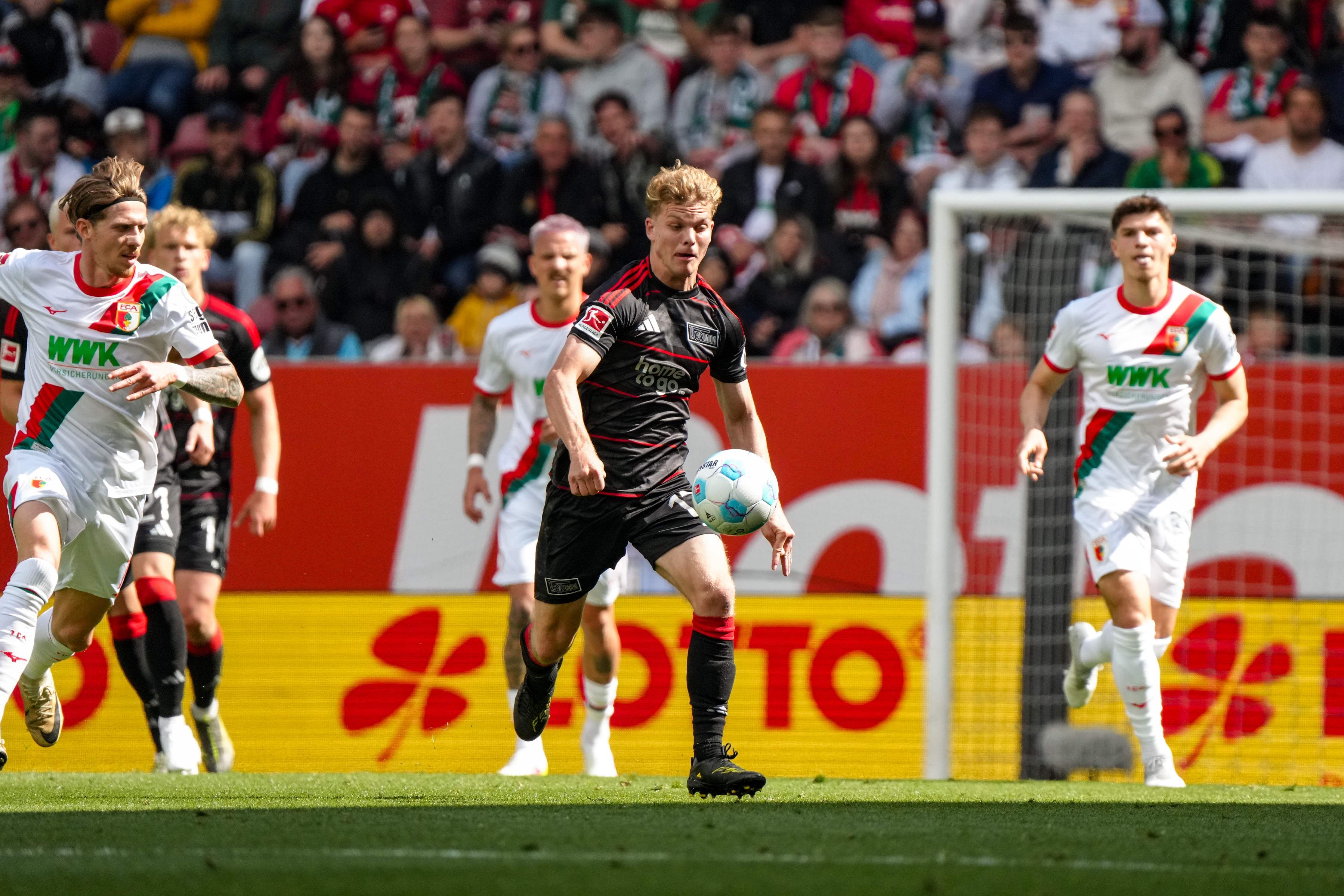 Ein Fußballspieler in schwarz-rot dribbelt mit dem Ball, während Spieler in weißen Trikots ihn verteidigen. Fans im Hintergrund.