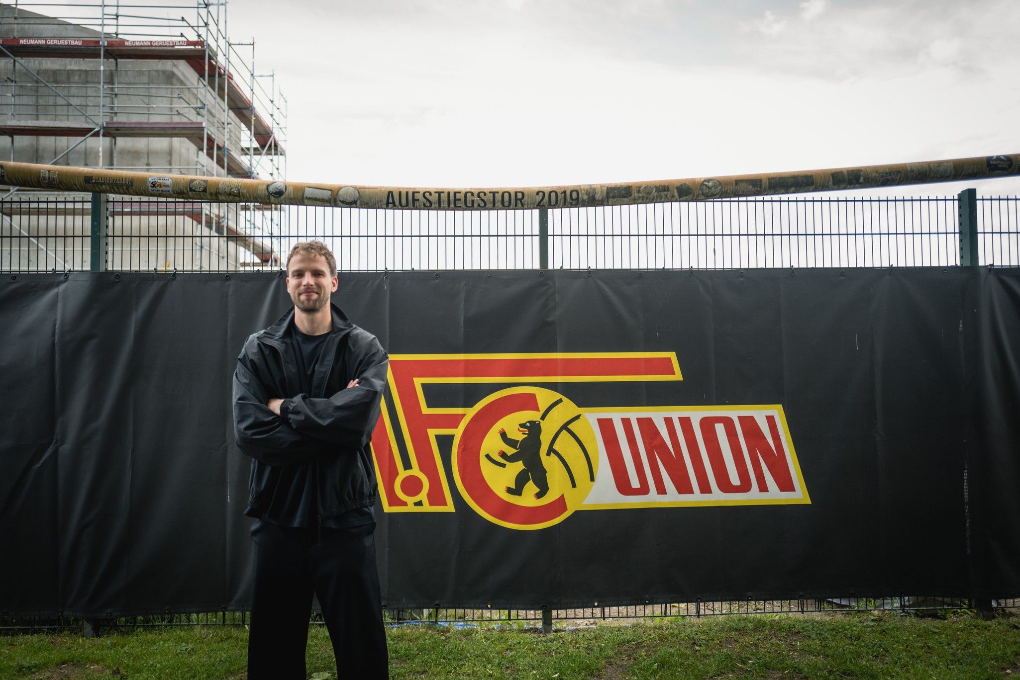 Ein Mann steht vor einem großen Banner mit dem Logo von FC Union. Im Hintergrund ist eine Baustelle zu sehen.