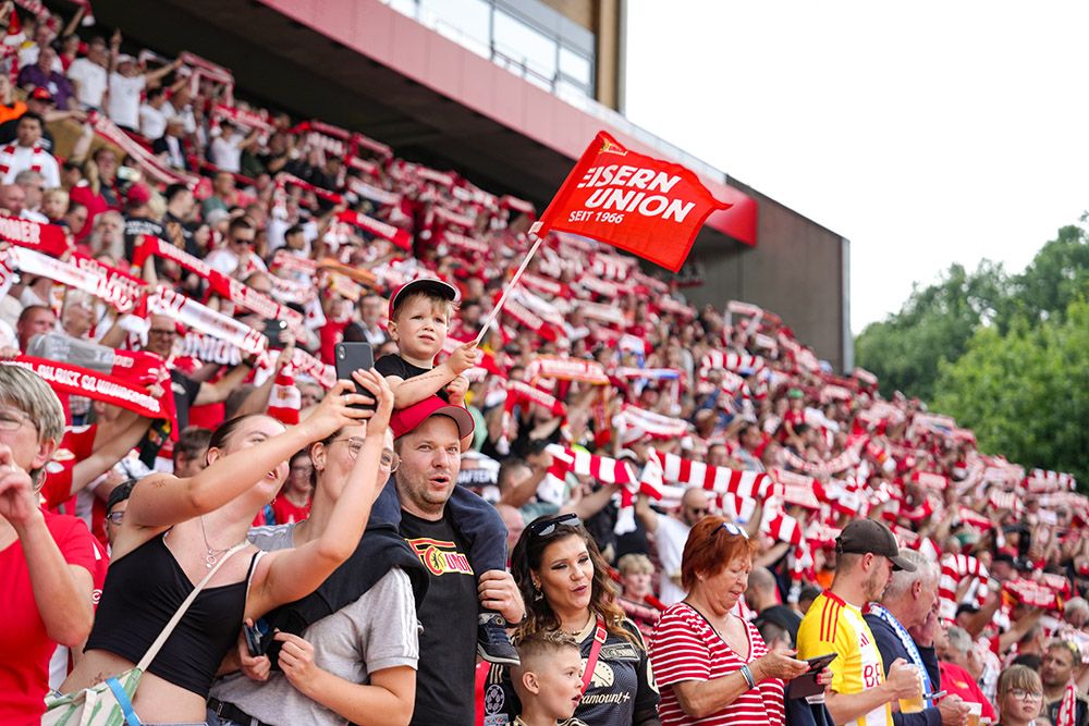 Ein begeistertes Publikum mit Fans, die Vereinsschals schwenken, darunter ein Kind mit einer Fahne, im Stadion.