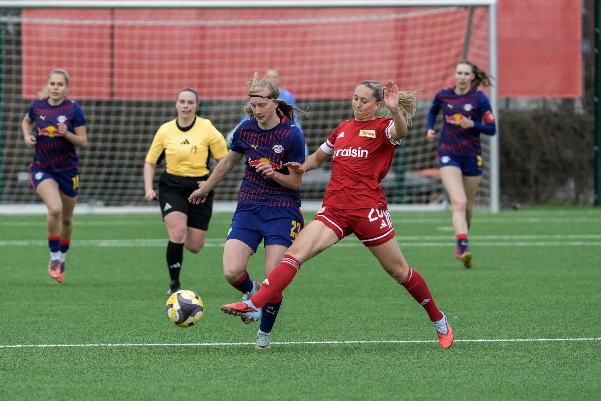 Zwei Frauenfußballspielerinnen kämpfen um den Ball auf einem Rasenfeld, im Hintergrund Schiedsrichterin und andere Spielerinnen.