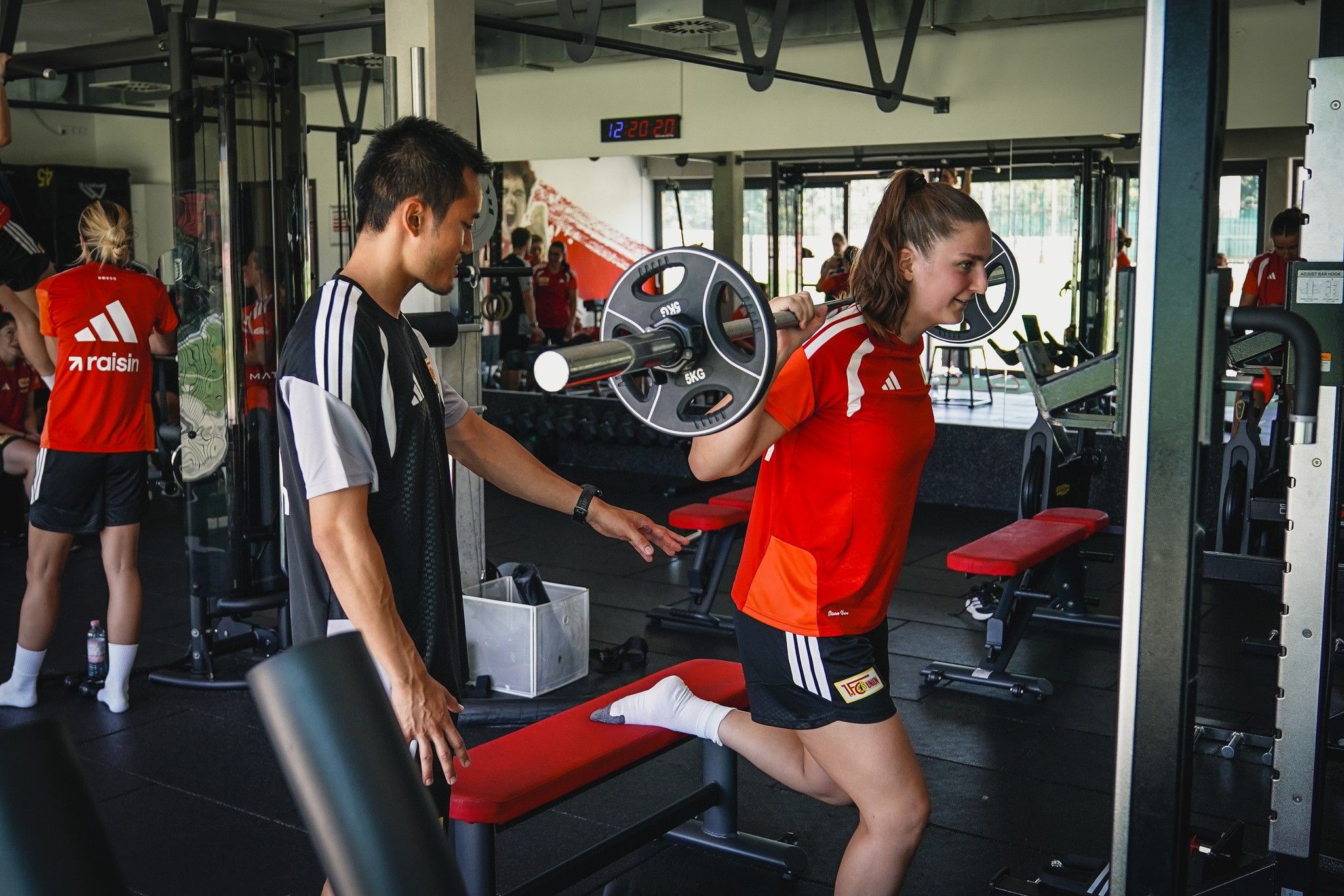 Eine Sportlerin in rotem Trikot trainiert mit einer Langhantel, während ein Trainer sie unterstützt. Im Hintergrund sind weitere Personen zu sehen.
