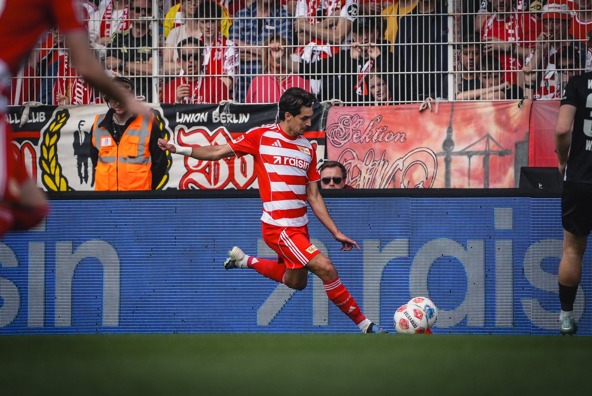 Spielszene im Fußball, ein Spieler in rot-weißer Uniform schießt den Ball, Zuschauer im Hintergrund.