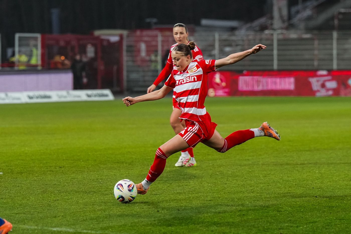 Spielerinnen in roten Fußballtrikots auf dem Platz; eine Spielerin tritt den Ball.
