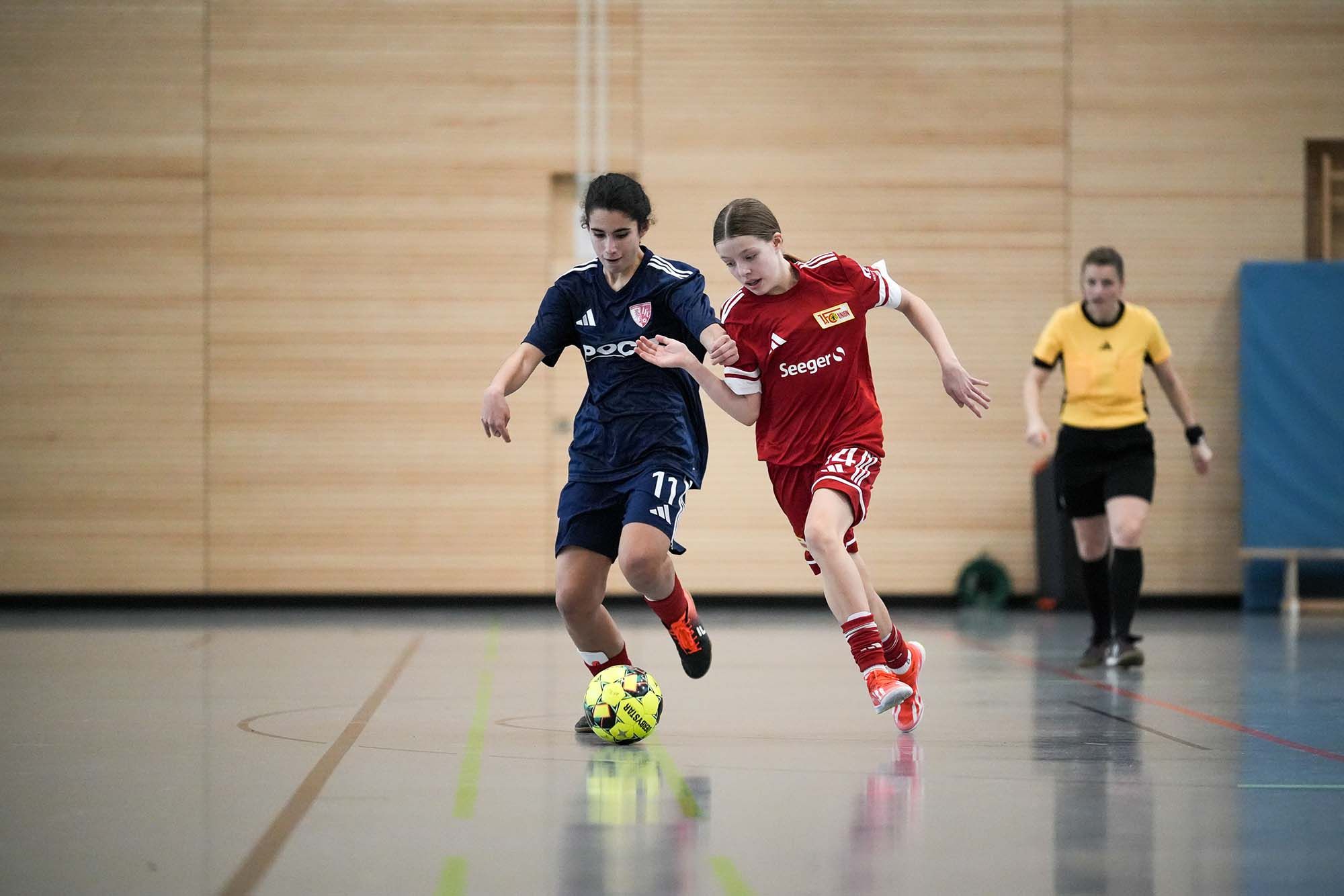 Zwei Mädchen spielen in einem Hallenfußballspiel. Eine trägt ein blaues, die andere ein rotes Trikot. Im Hintergrund eine Schiedsrichterin.
