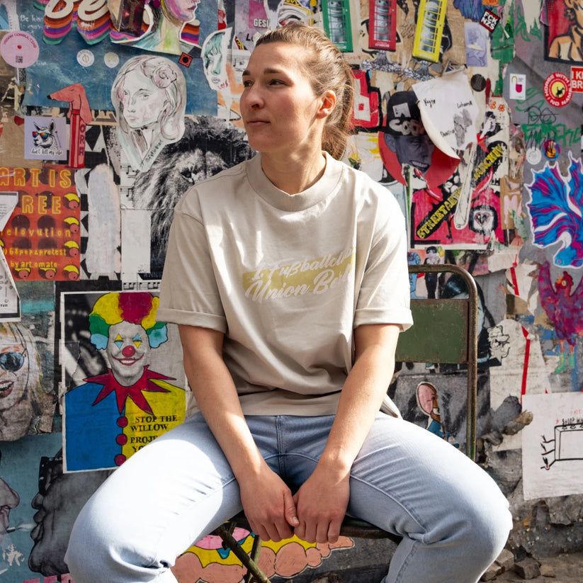 Frau sitzt auf einem Stuhl vor einer bunten Wand mit Plakaten und Street Art, trägt ein beiges T-Shirt und Jeans.
