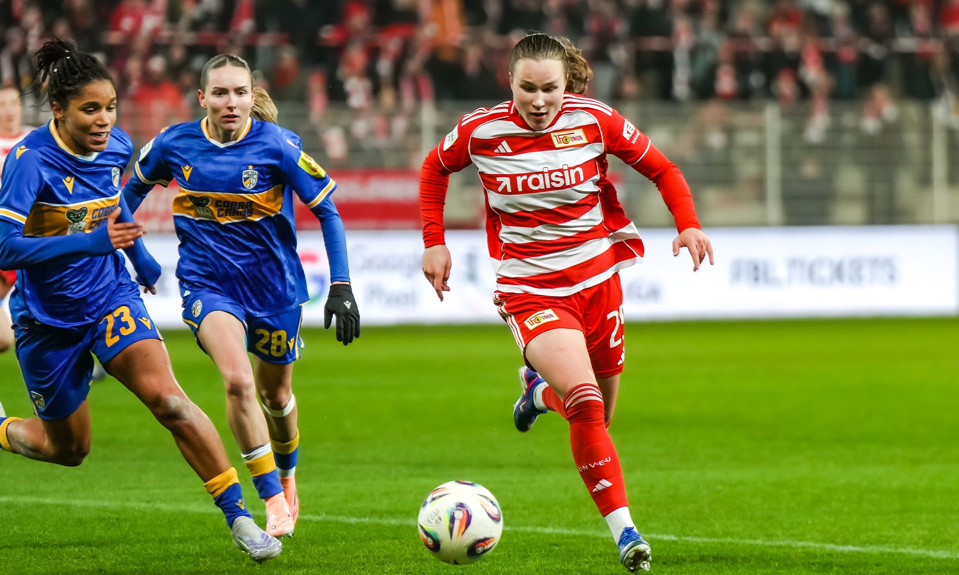 Eine Spielerin in roten Trikots sprintet mit dem Ball, während zwei Gegnerinnen in blauen Trikots sie verfolgen.