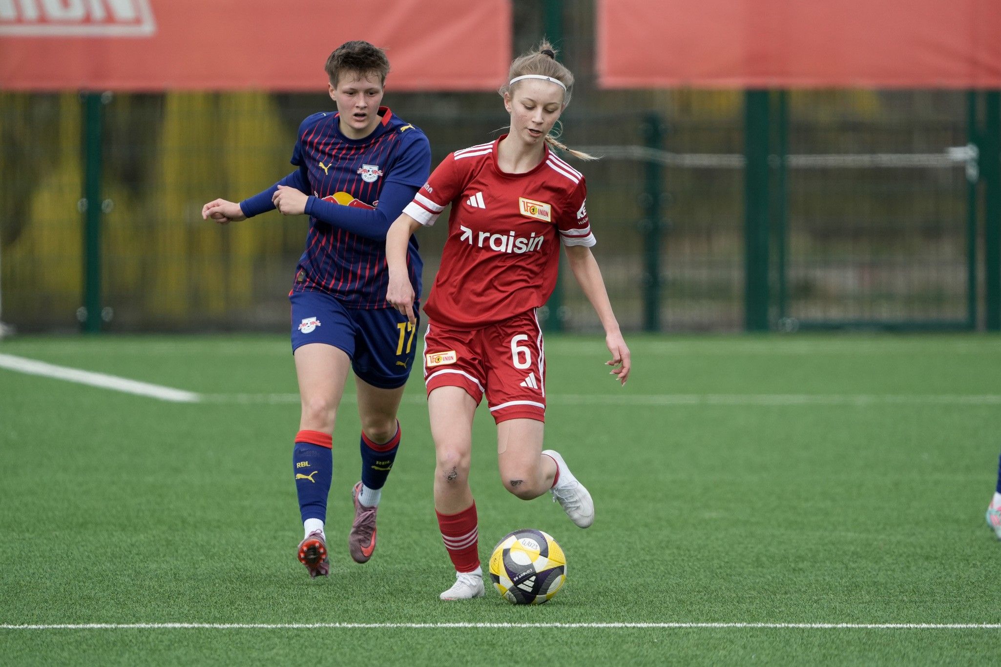 Zwei Frauenfußballspielerinnen auf einem Spielfeld, eine in rotem Trikot mit Ball, die andere verfolgt sie.