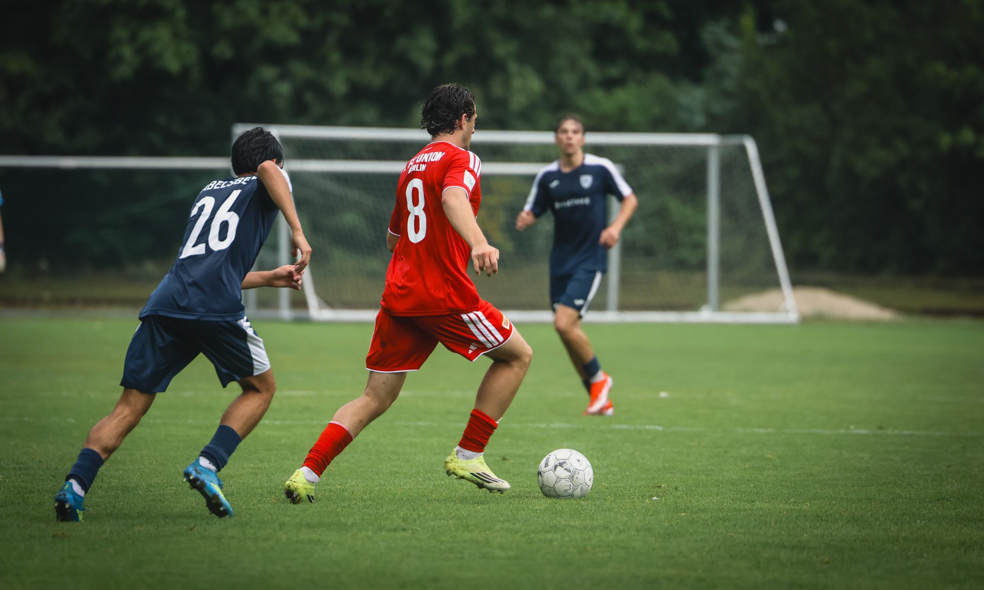 Ein roter Spieler mit der Nummer 8 dribbelt den Ball, während ein blauer Spieler mit der Nummer 26 ihn verfolgt. Im Hintergrund ein Fußballtor.