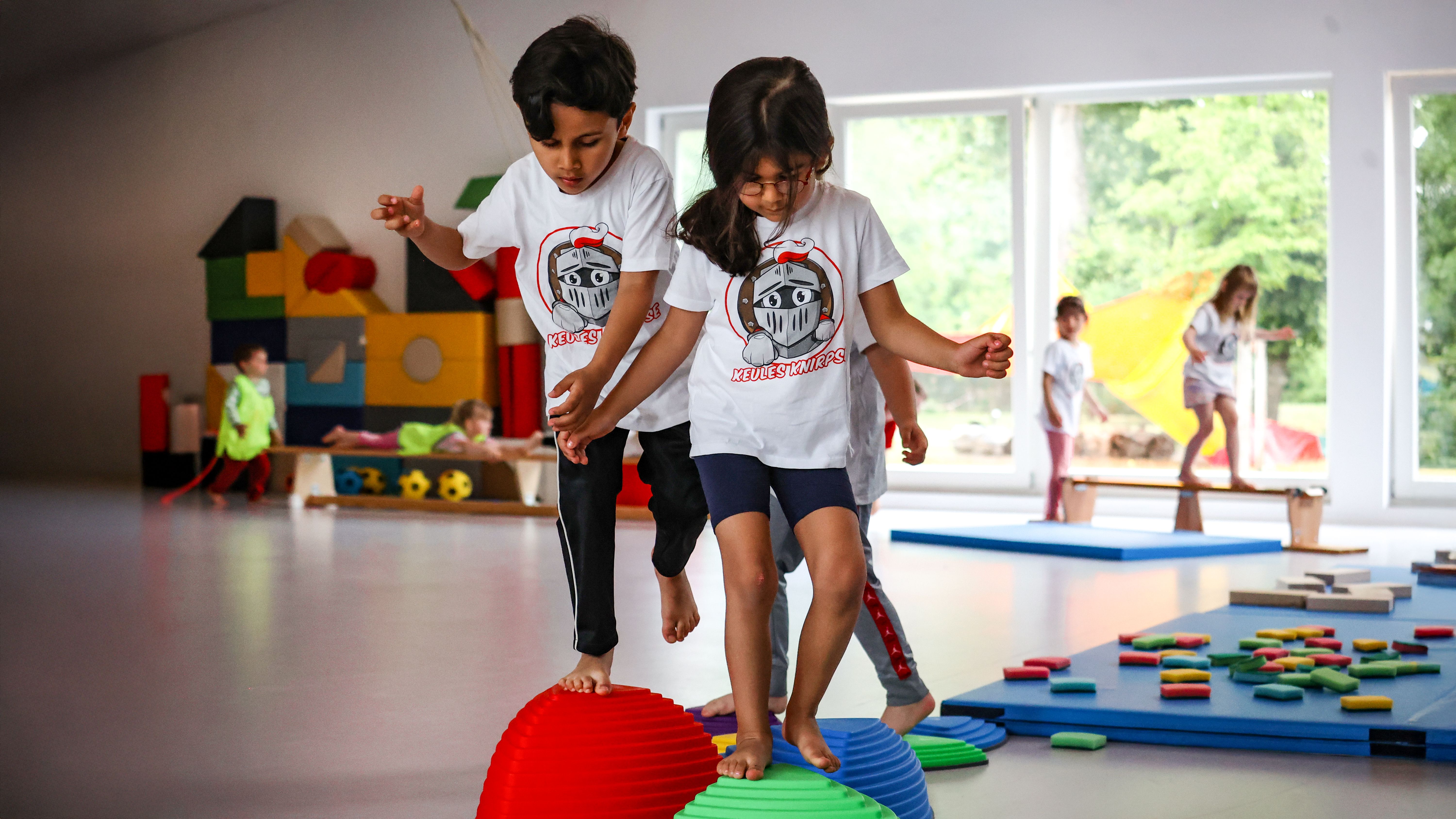 Zwei Kinder balancieren auf bunten Kletterskulpturen in einem Spielzimmer. Im Hintergrund sind weitere Kinder aktiv.
