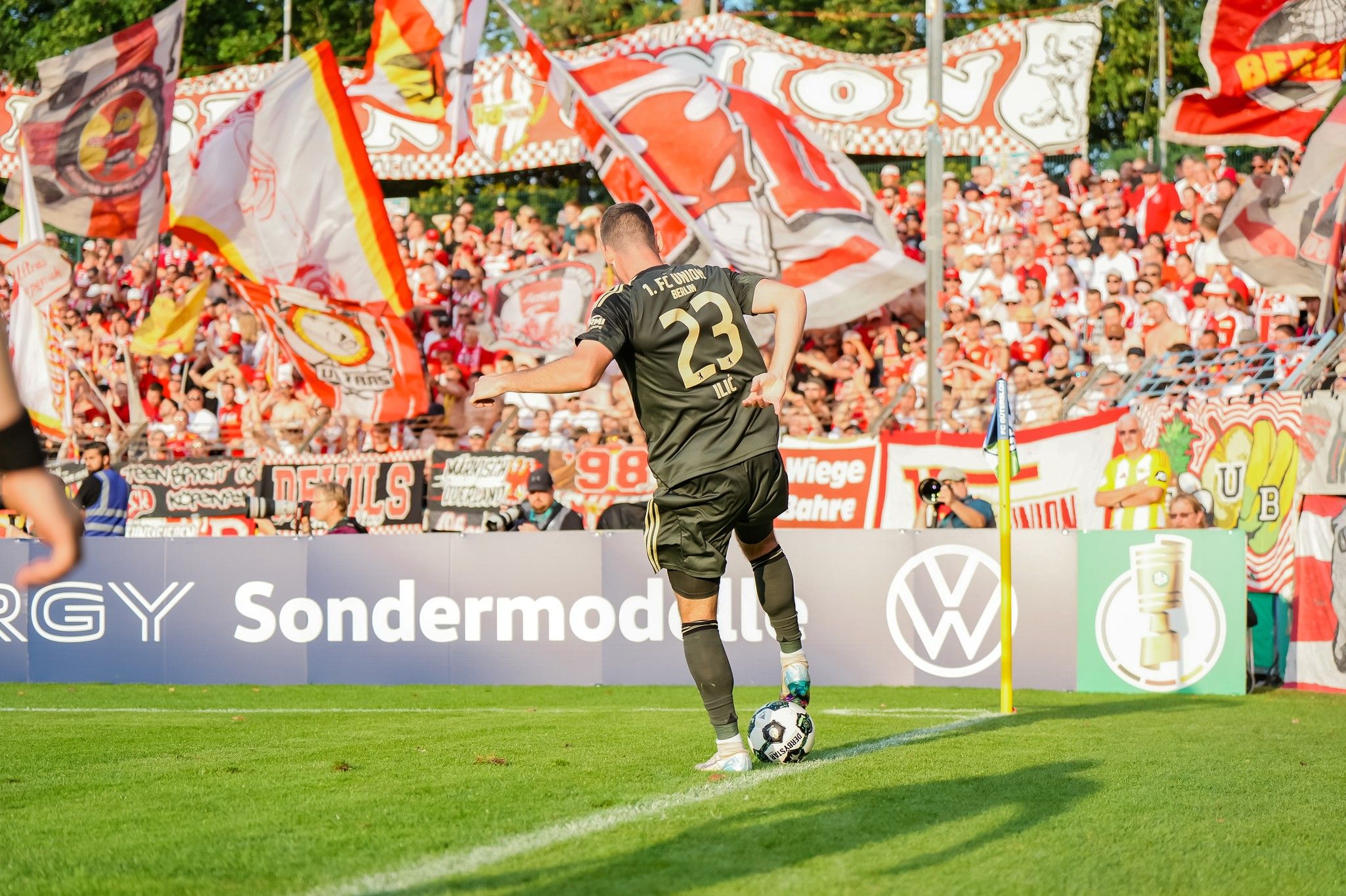 A goalkeeper in black gear is preparing for a corner kick, while enthusiastic fans wave flags in the background.