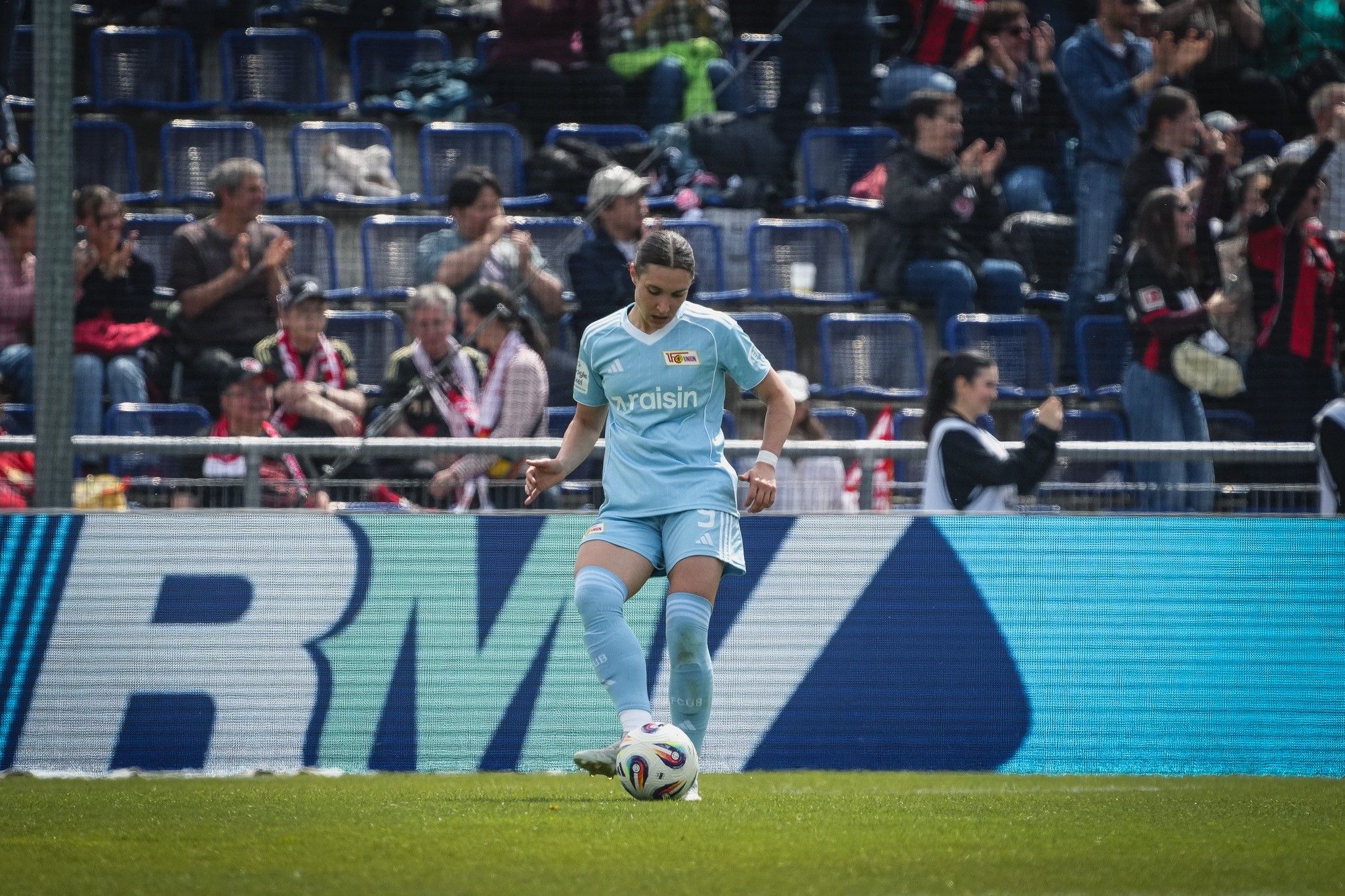 Spielerin in hellblauer Fußballausrüstung bereitet sich darauf vor, einen Ball auf dem Spielfeld zu spielen. Zuschauer im Hintergrund.