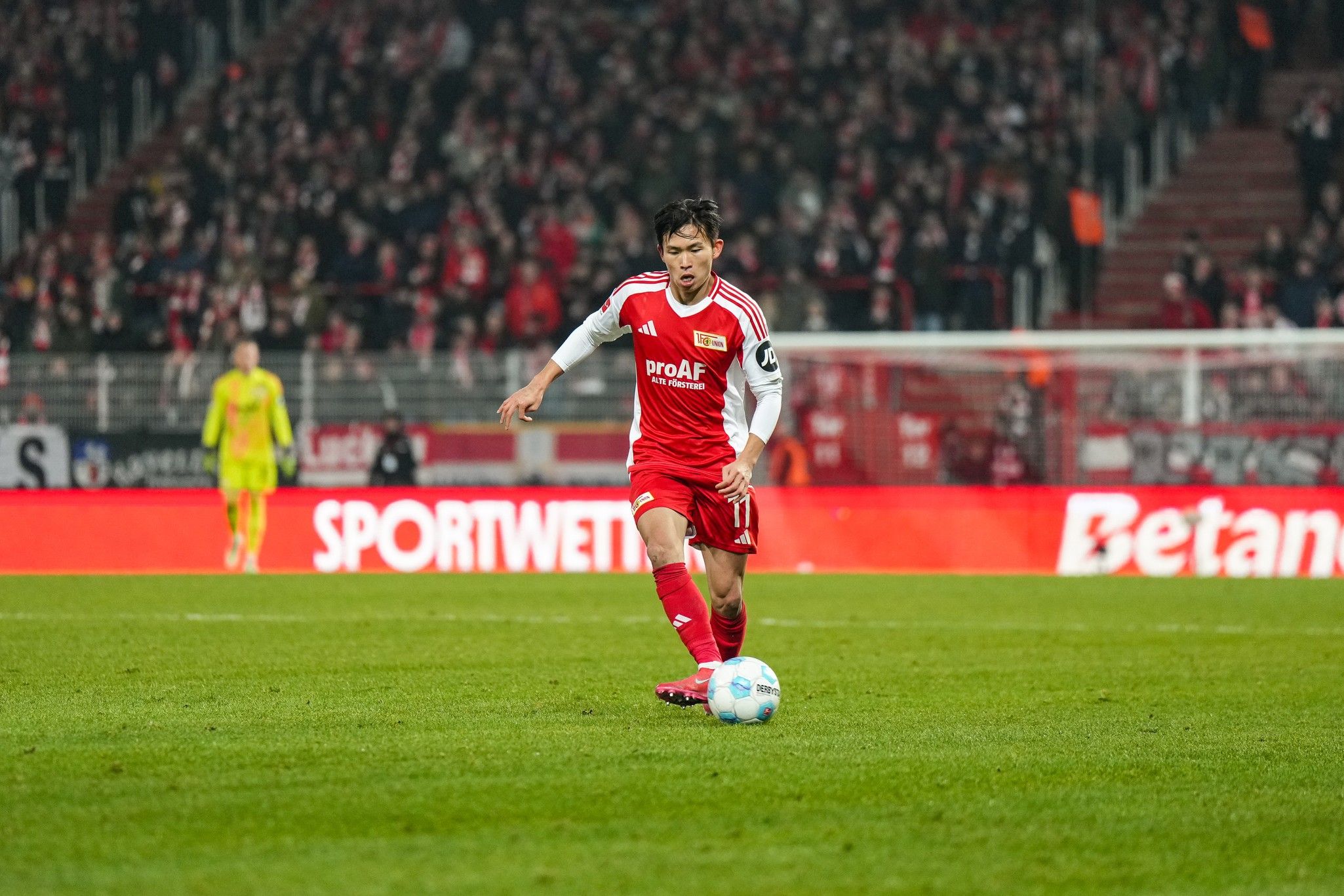 Spieler in rotem Trikot dribbelt den Ball auf dem Spielfeld, im Hintergrund jubelnde Fans und eine Sportarena.