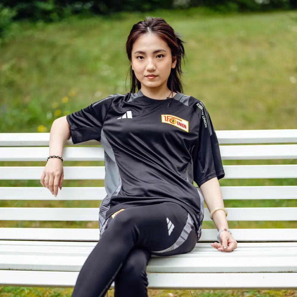 Eine Person sitzt auf einer Bank im Freien und trägt ein schwarzes Sporttrikot und Leggings.