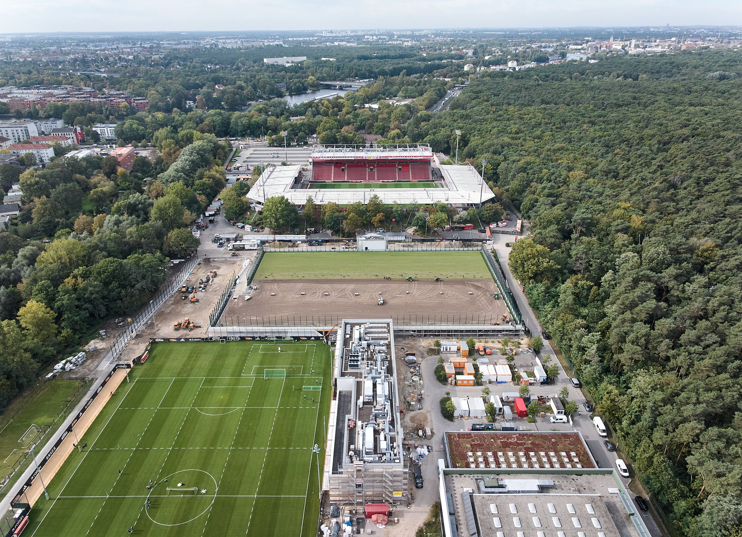 Luftaufnahme eines Sportgeländes mit Fußballfeldern, einem Stadion und umliegendem Wald.
