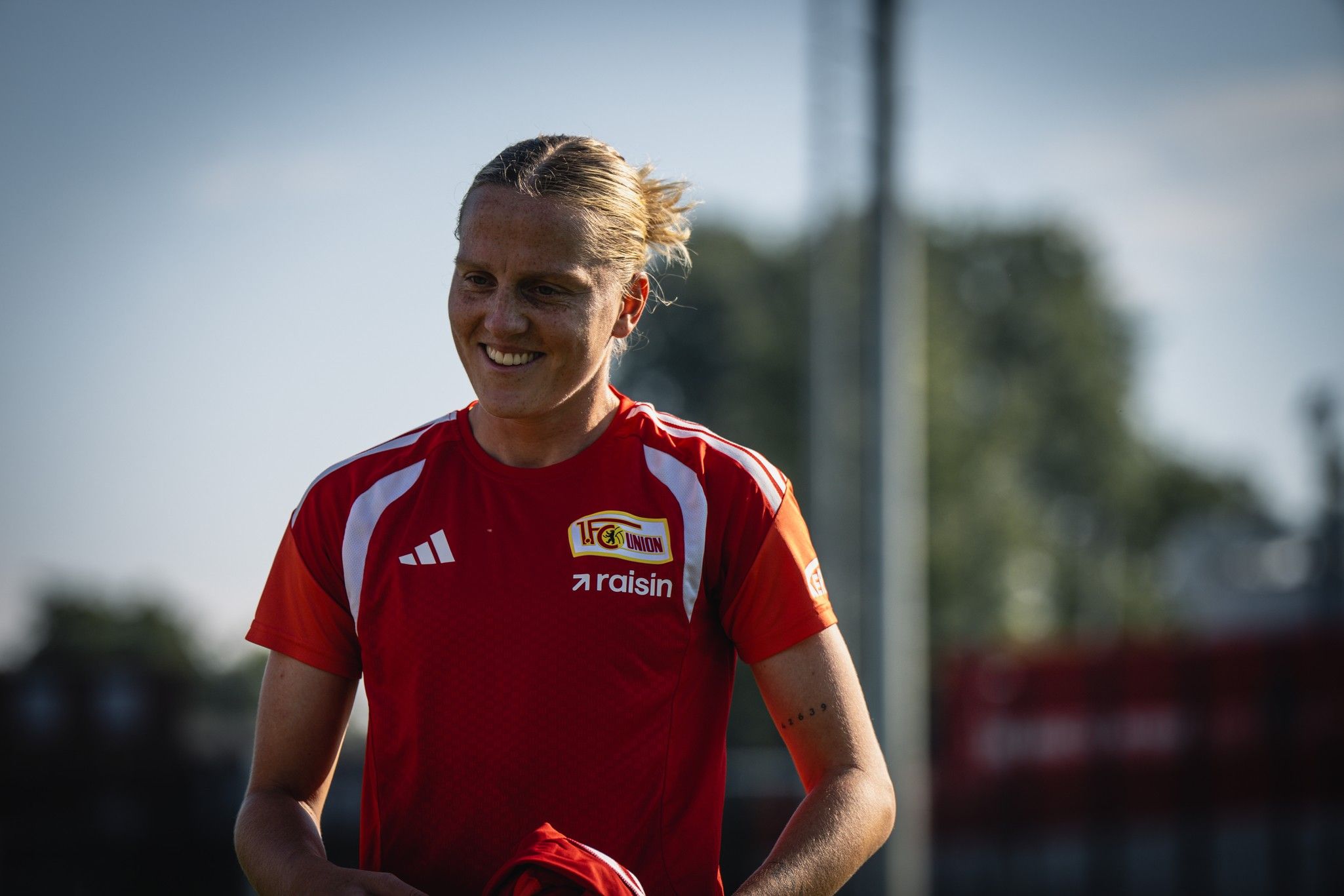 Eine lachende Person trägt ein rotes Sporttrikot mit Vereinslogo und hält ein rotes Kleidungsstück in der Hand.