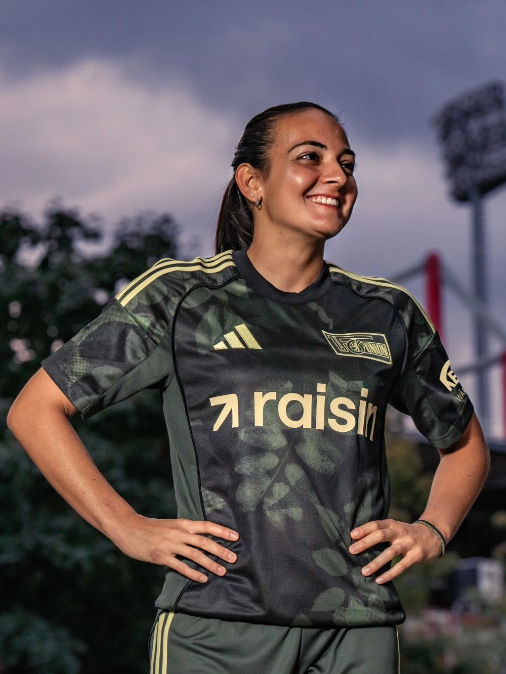 Frau in einem sportlichen Trikot mit camouflage-muster und Aufschrift "raisin", lächelt vor einem Fußballstadion.