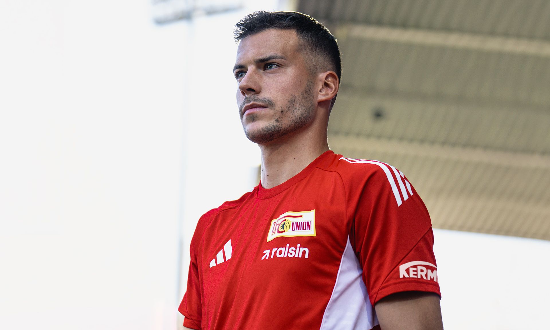 Ein Fußballspieler in rot-weißer Vereinskleidung steht auf einem Spielfeld, schaut nachdenklich und pose für ein Foto.