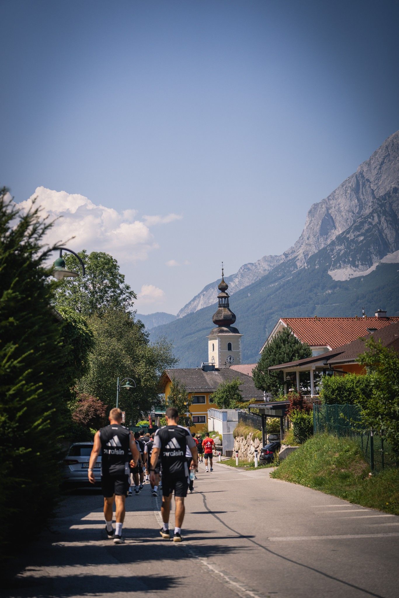 Zwei Personen gehen eine Straße entlang, im Hintergrund ist eine Kirche und die Berge zu sehen. Sonniger Tag mit blauem Himmel.