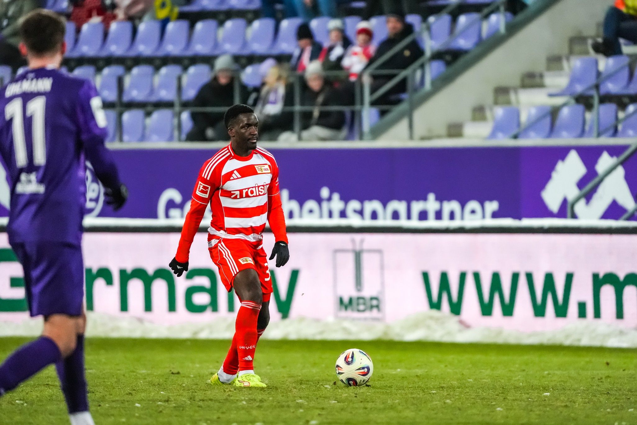 Ein Fußballspieler in roter und weißer Uniform steht auf dem Spielfeld, während ein gegnerischer Spieler in lila hinter ihm steht.