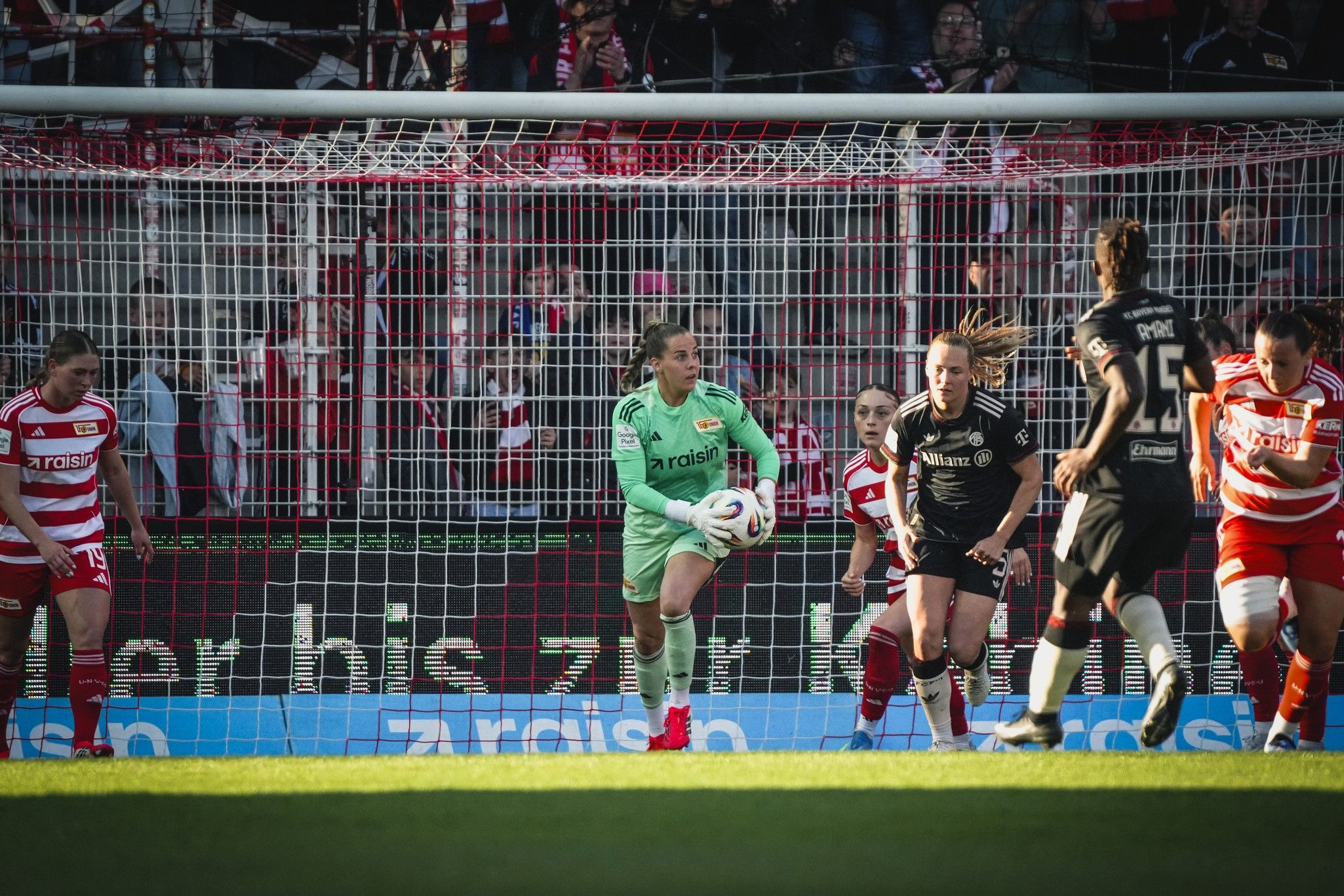 Torhüterin in grüner Ausrüstung fängt den Ball im Spiel, umgeben von spielenden Gegnerinnen und einer jubelnden Menge im Hintergrund.