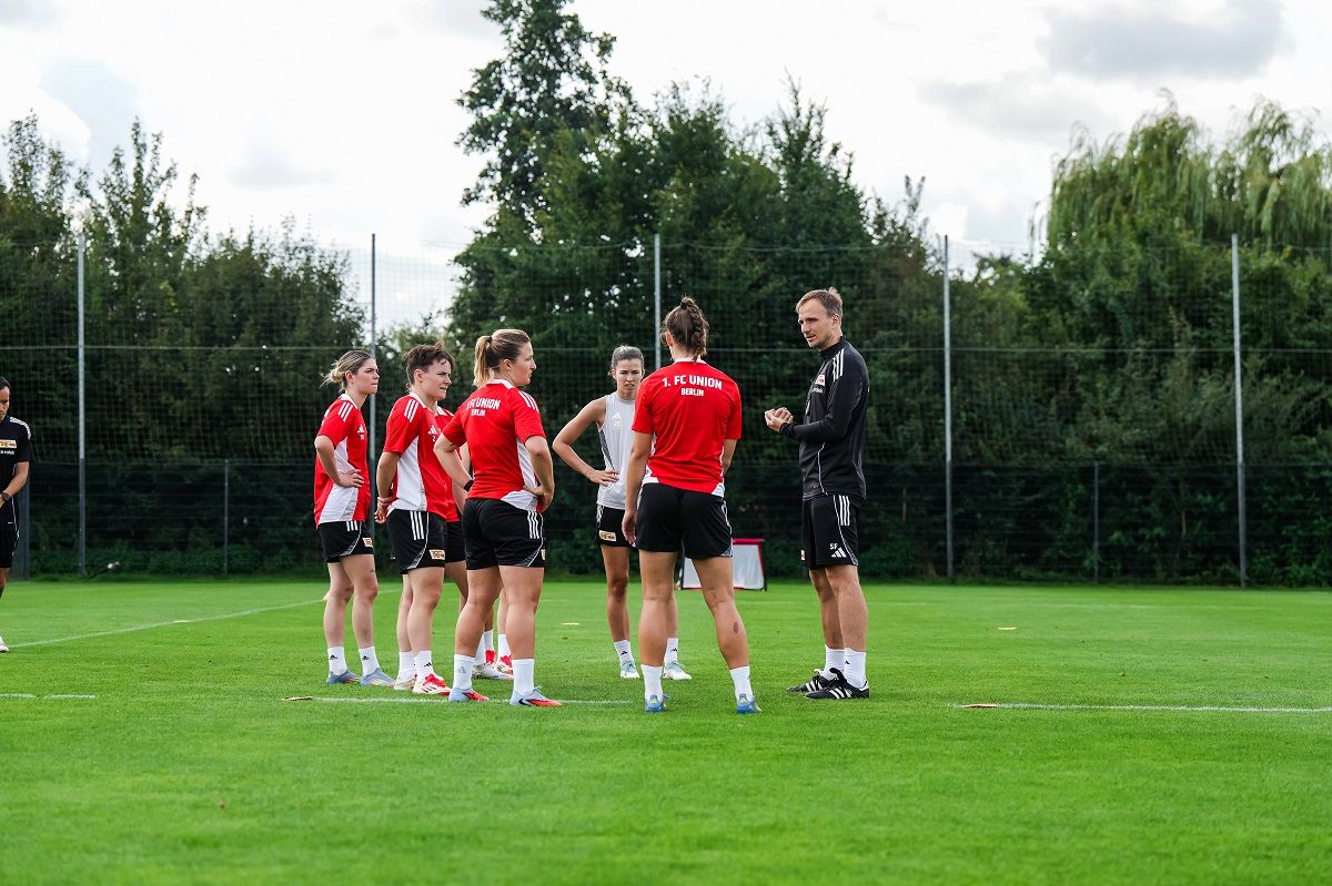 Eine Gruppe von Fußballspielerinnen in roten Trikots steht auf einem Trainingsplatz und hört einem Trainer zu.