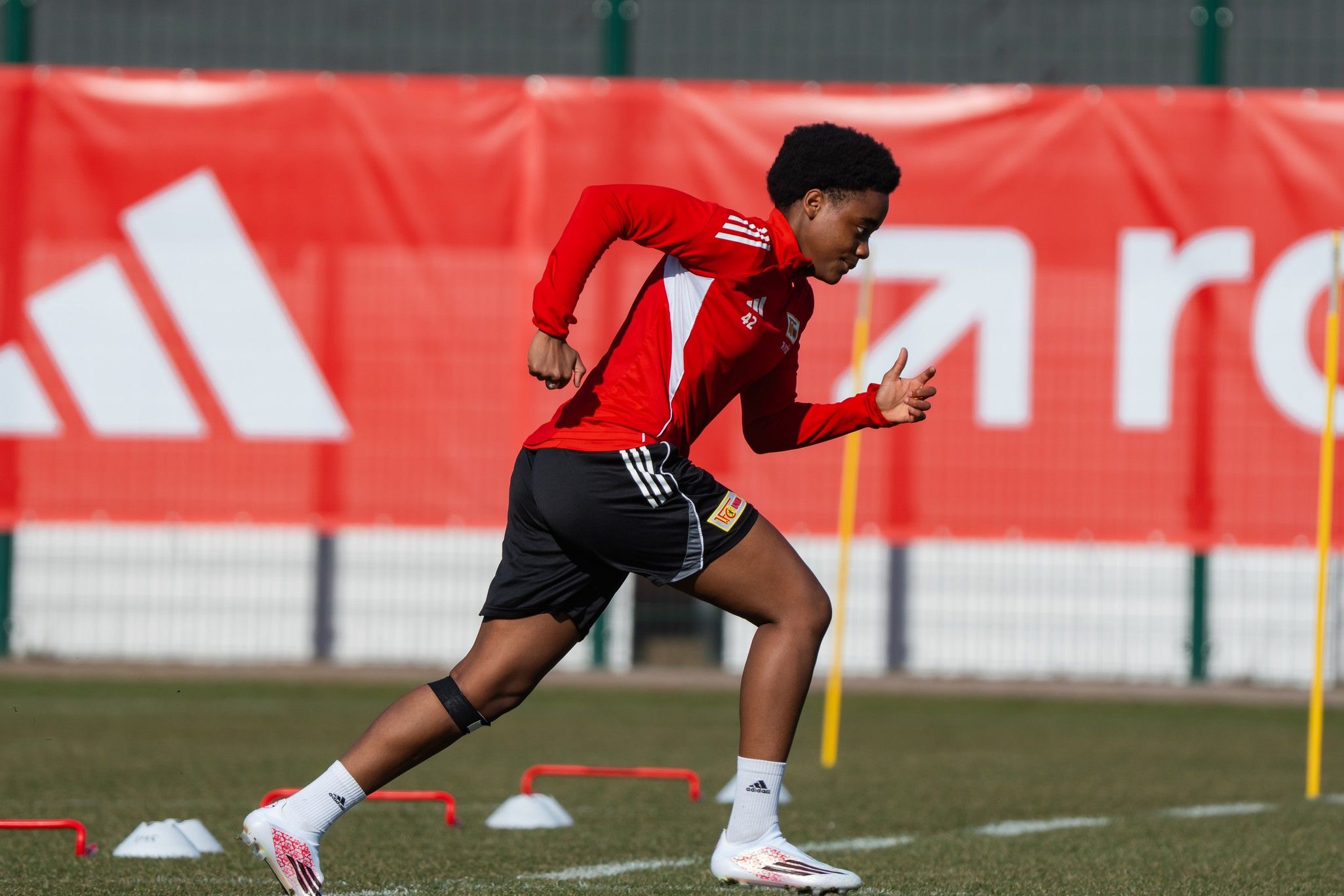 Ein Sportler in rotem Trikot sprintet während des Trainings auf einem Fußballfeld, umgeben von Trainingsgeräten und einer Werbebande.