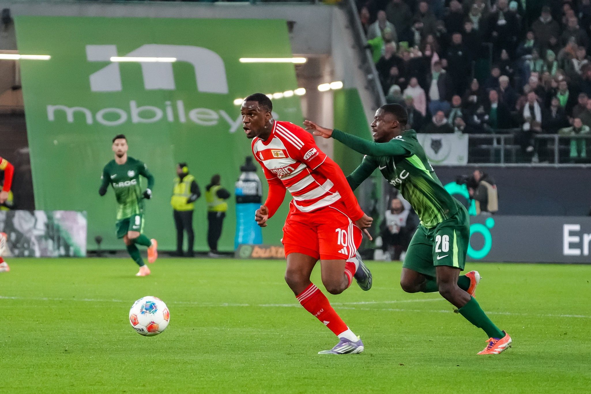 Ein Spieler in rotem Trikot sprintet auf einem Fußballfeld, während ein Verteidiger in grünem Trikot ihn verfolgt.