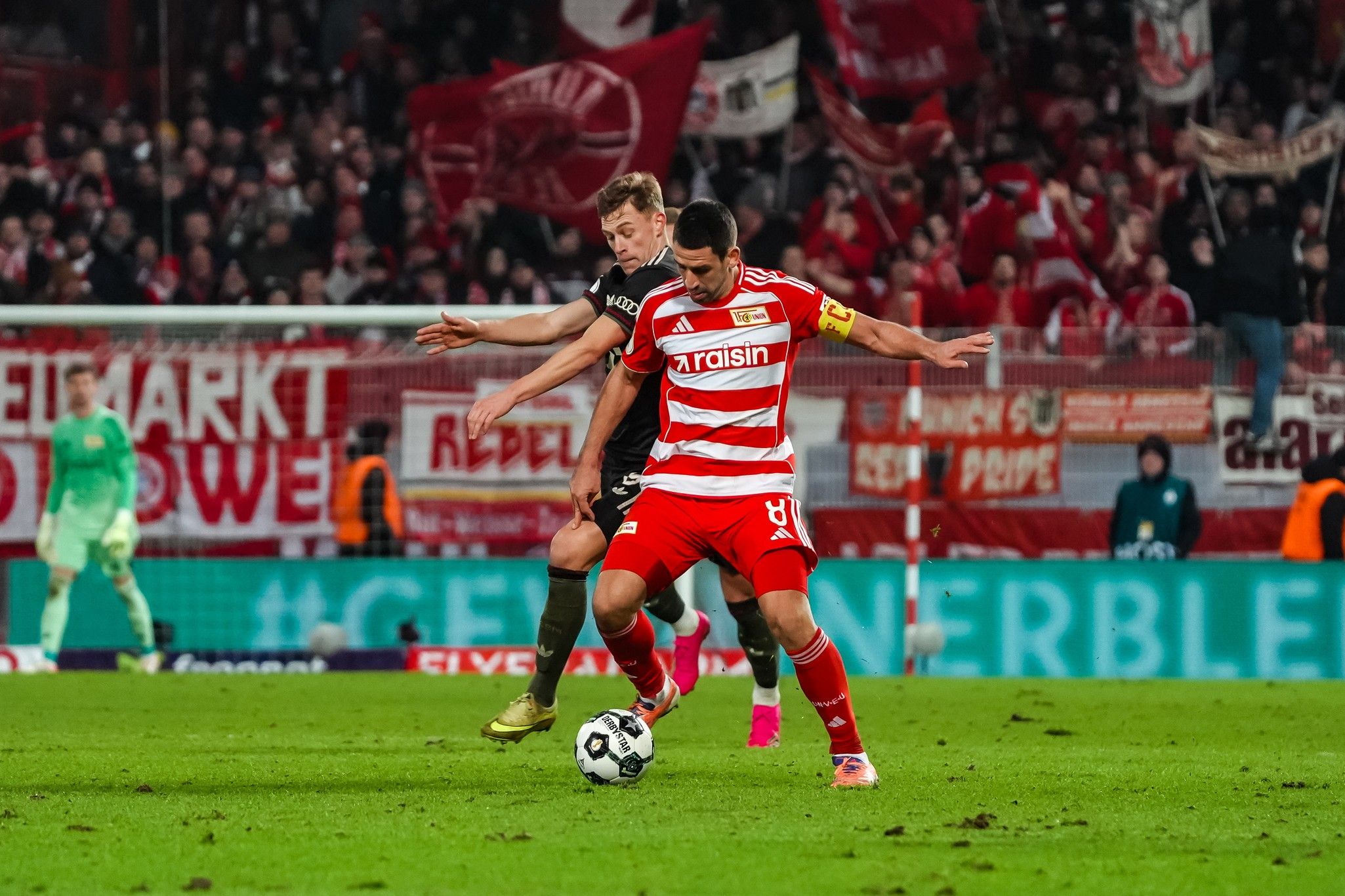 Zwei Fußballspieler im Wettkampf um den Ball, mit Fans und Fahnen im Hintergrund.