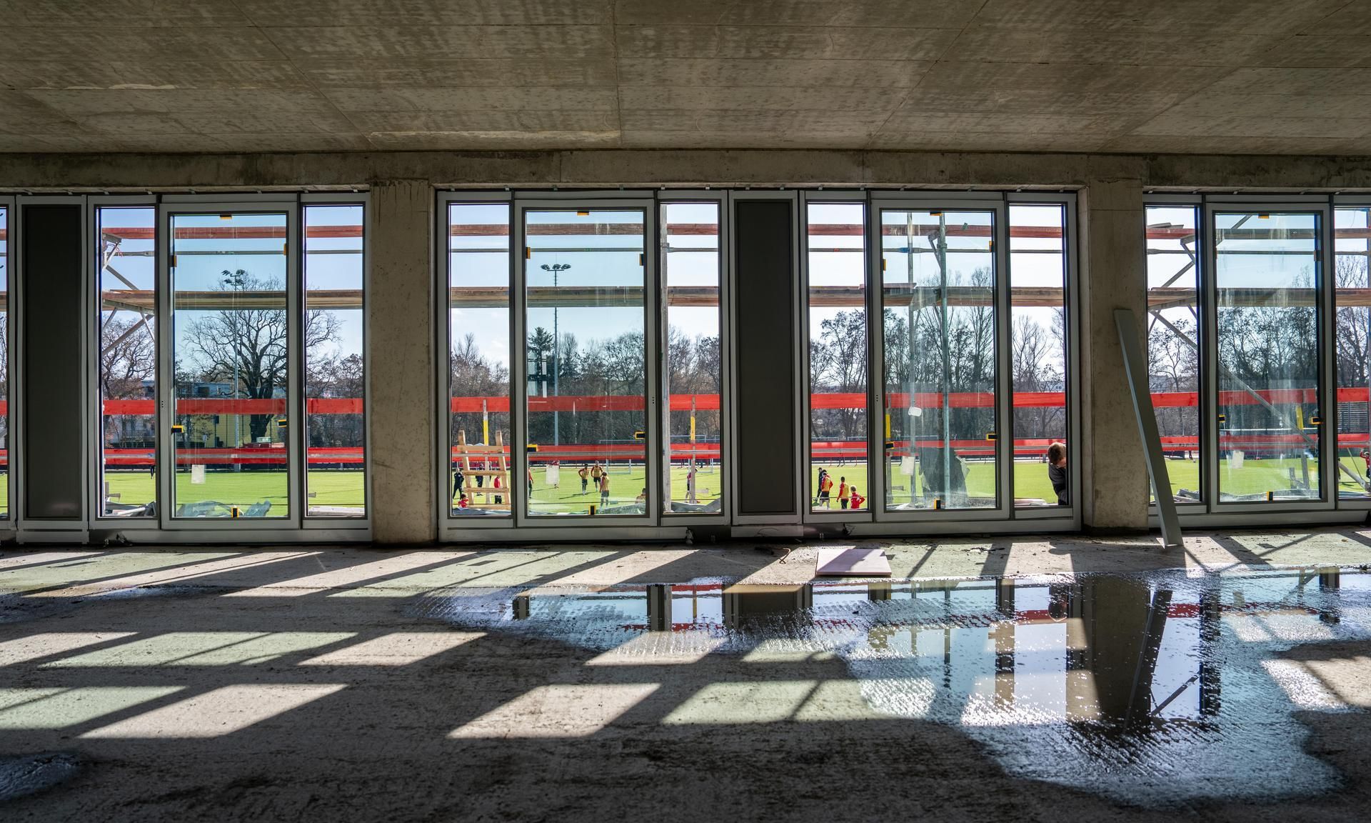 Blick durch große Fenster auf ein sportliches Außengelände, der Boden ist nass und teilweise von Licht- und Schattenmustern durchzogen.