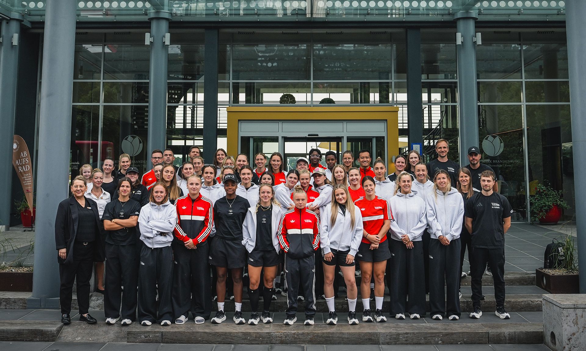 Gruppenfoto von Männern und Frauen in Trainingskleidung, die vor einem modernen Gebäude stehen.