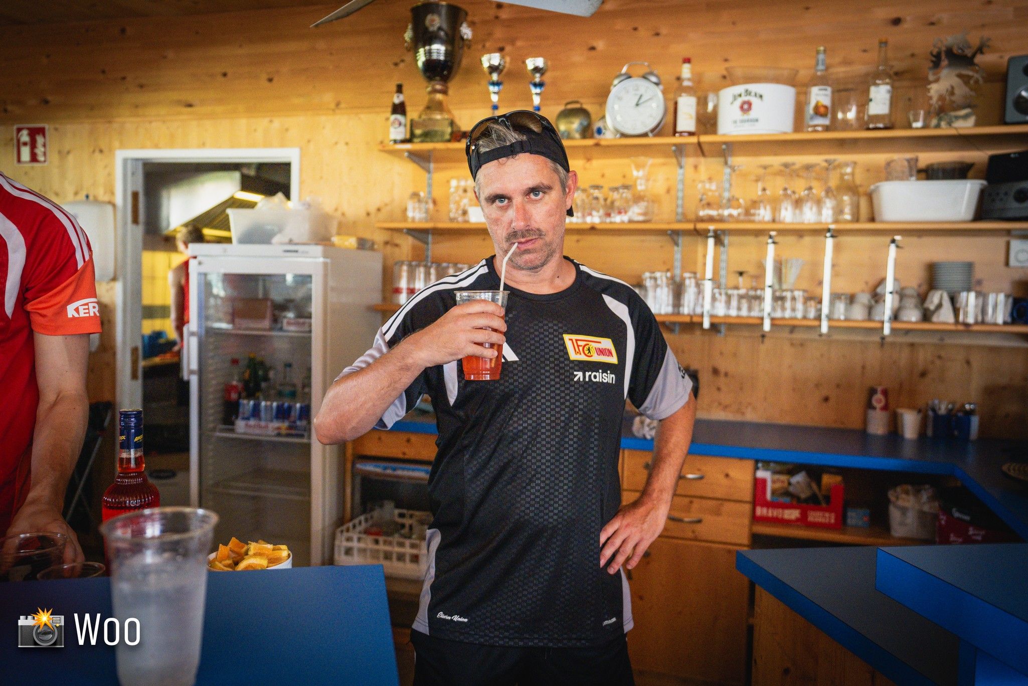 Ein Mann in einem sportlichen Outfit steht an einer Bar und hält ein Getränk in der Hand. Im Hintergrund stehen Gläser und Spirituosen.