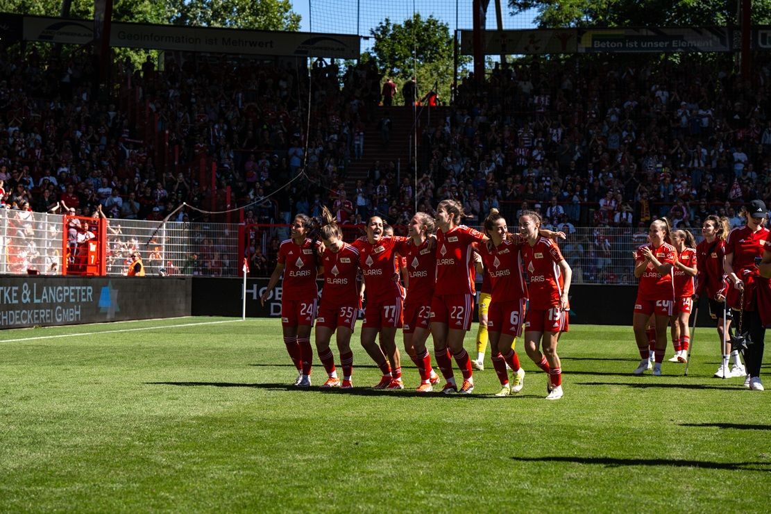 Eine Gruppe von Fußballspielern in roten Trikots feiert auf einem Rasenfeld vor jubelnden Fans.