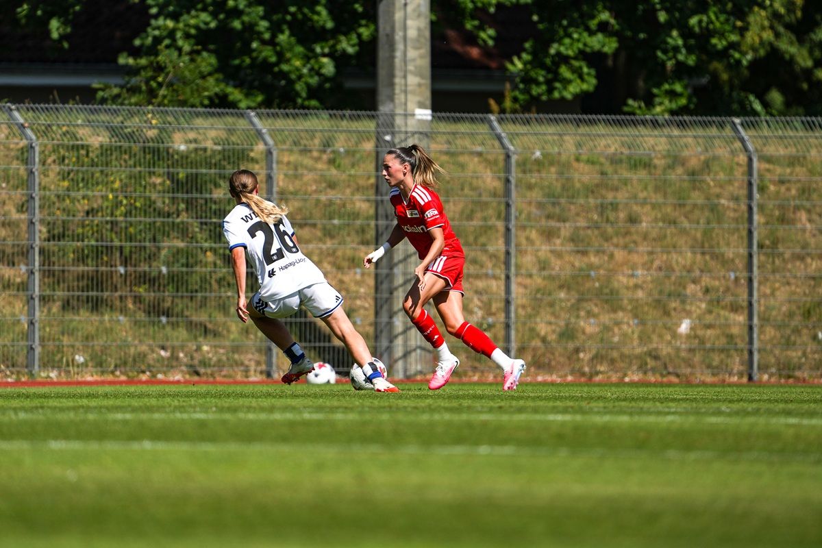 Spielszene im Fußball: Eine Spielerin im roten Trikot verfolgt eine Gegnerin im weißen Trikot auf einem Rasenfeld.