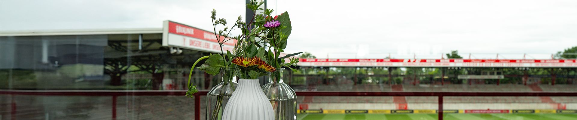 Vase mit Blumen auf einem Tisch, im Hintergrund ein unbesetztes Stadion mit grüner Rasenfläche.