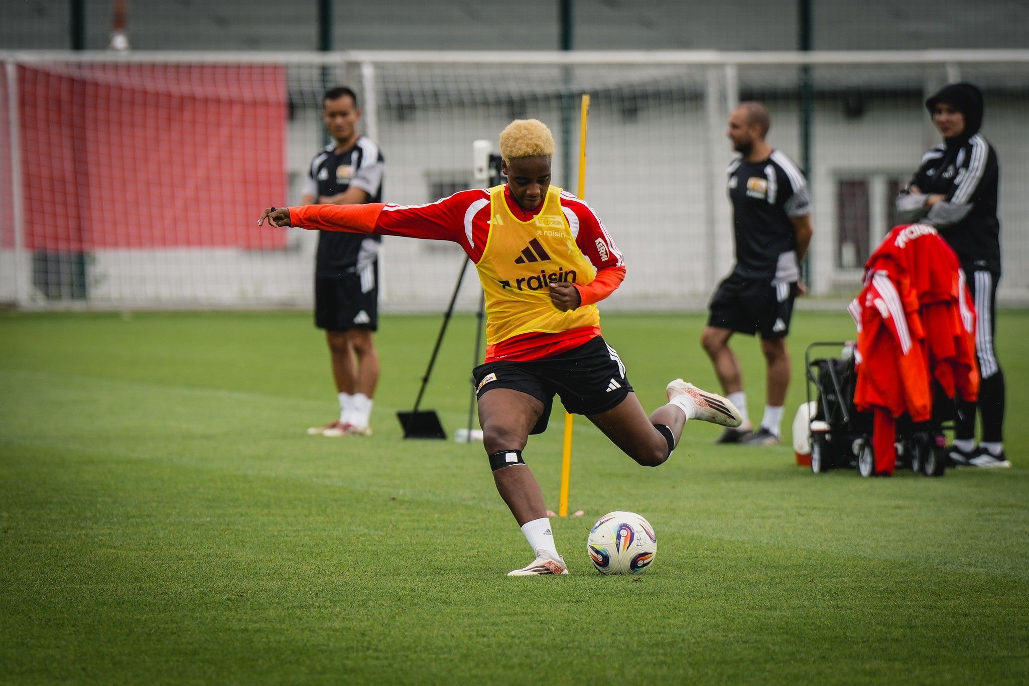 Eine Fußballspielerin mit blonden Haaren trainiert auf dem Platz, während Teamkollegen im Hintergrund zuschauen.