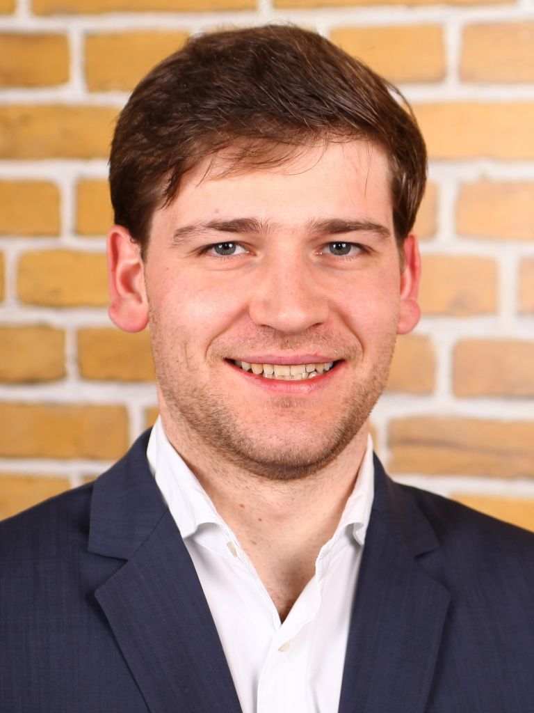 Ein junger Mann mit kurzen, braunen Haaren, lächelt in einem schwarzen Anzug vor einer Backsteinmauer.