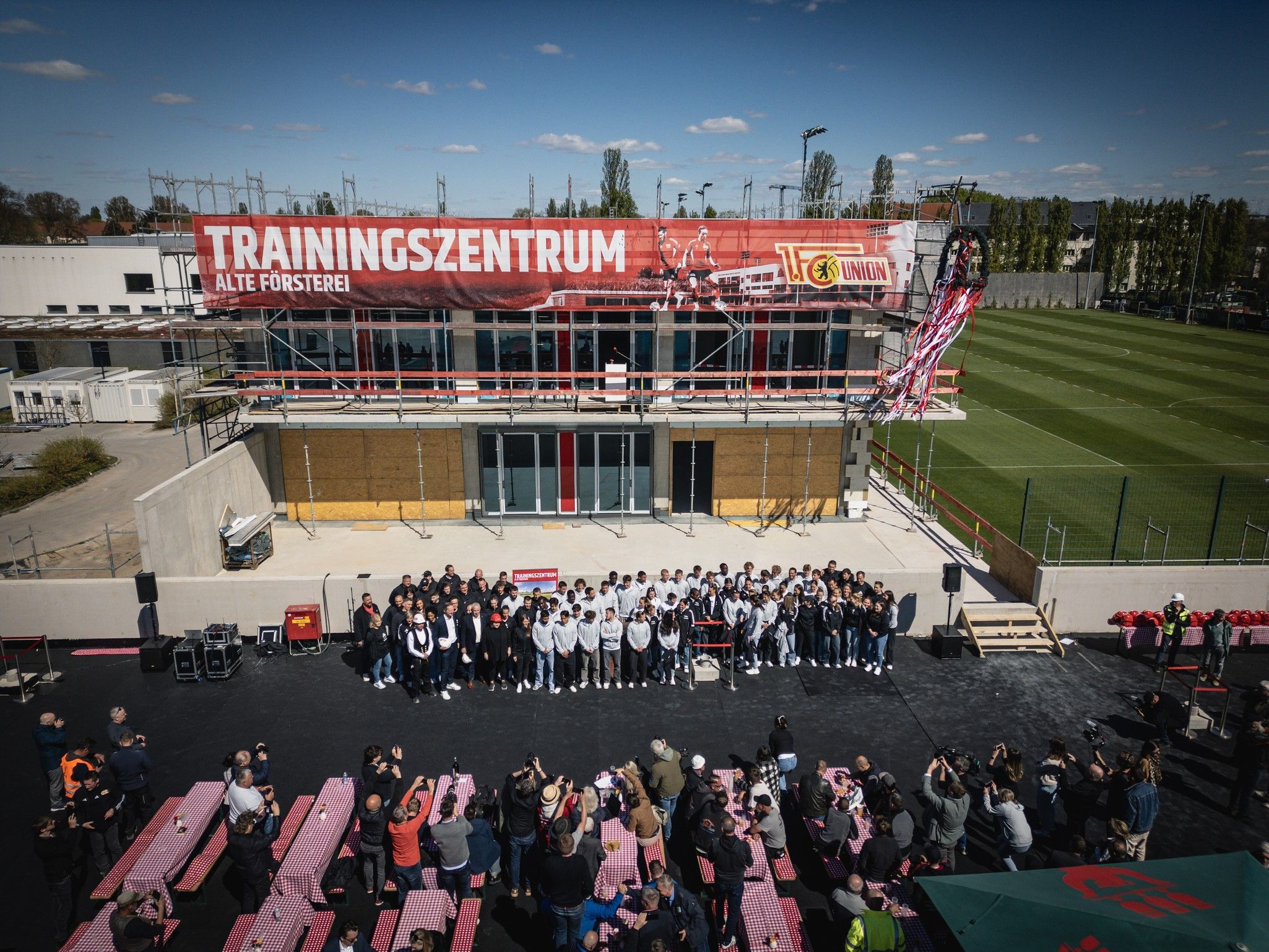 Gruppenfoto einer Mannschaft vor einem neuen Trainingszentrum, mit Zuschauern und einer großen Bannerbeschriftung im Hintergrund.