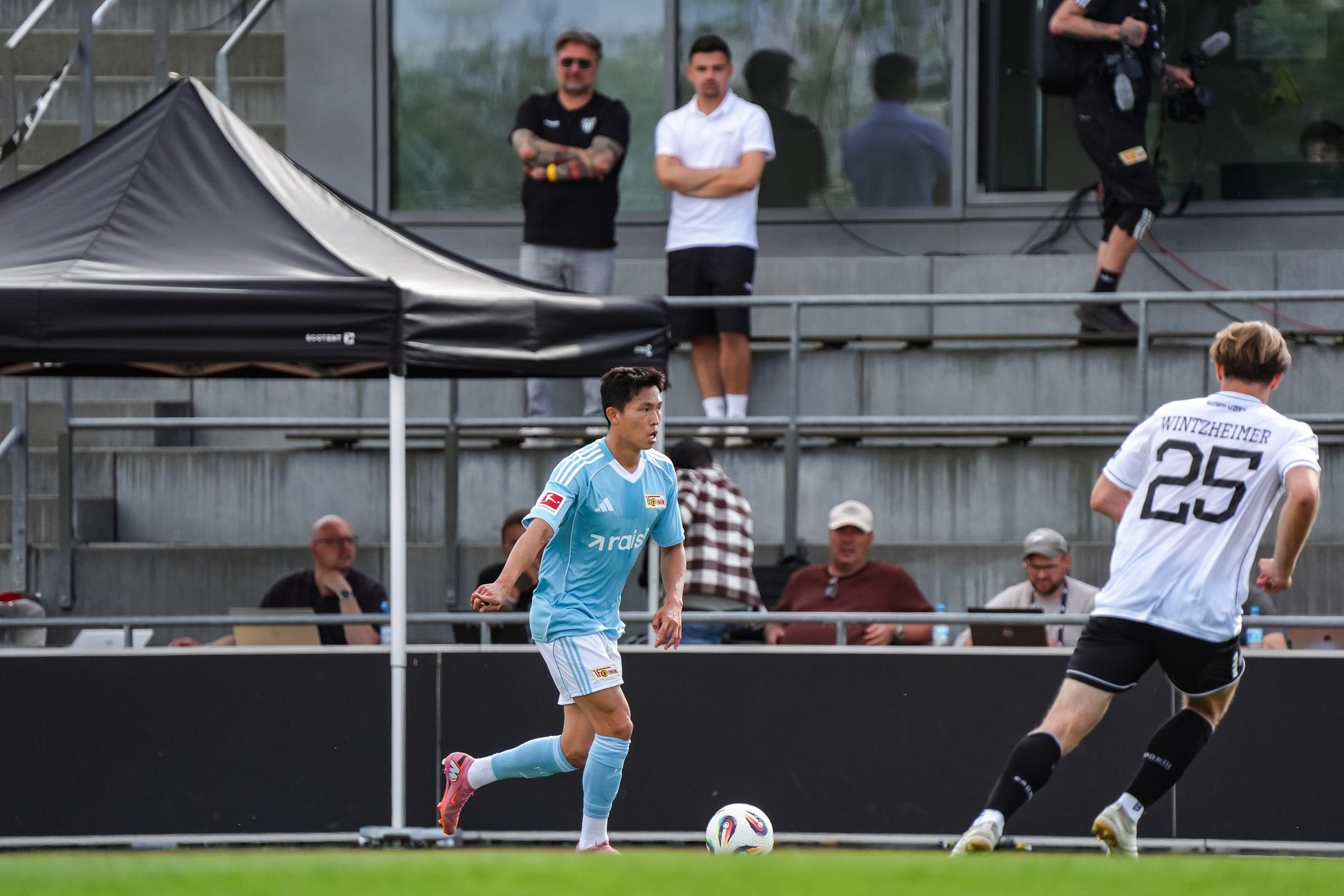 Ein Fußballspieler in hellblauer Kleidung dribbelt den Ball, während Zuschauer und Kamerateams im Hintergrund zuschauen.