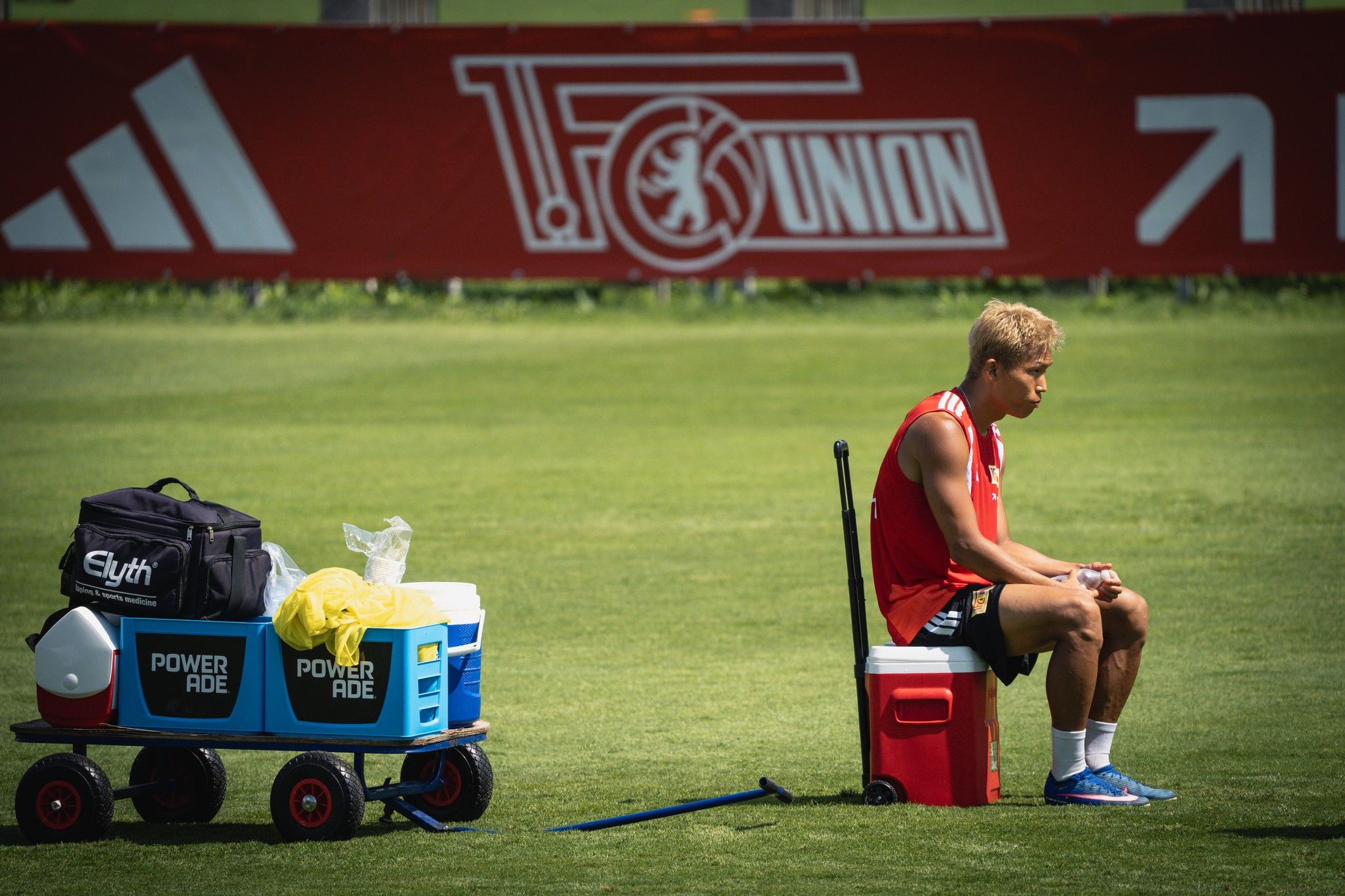 Ein junger Sportler sitzt auf einer Bank neben einem Kühler und einer Transportbox auf einem Fußballfeld.