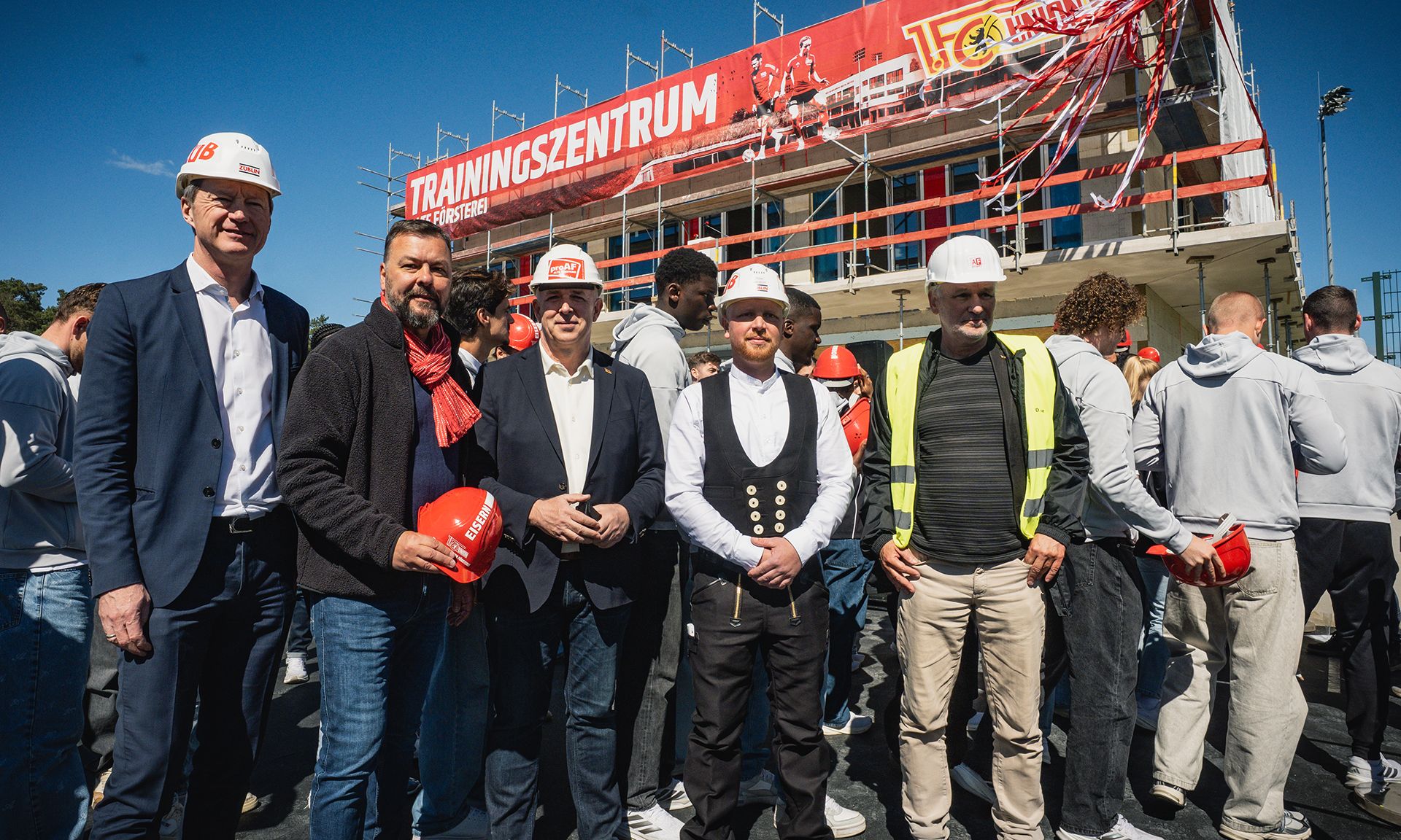 Gruppierung von fünf Männern in Schutzhelmen vor einem neuen Trainingszentrum mit Bauarbeitern im Hintergrund.