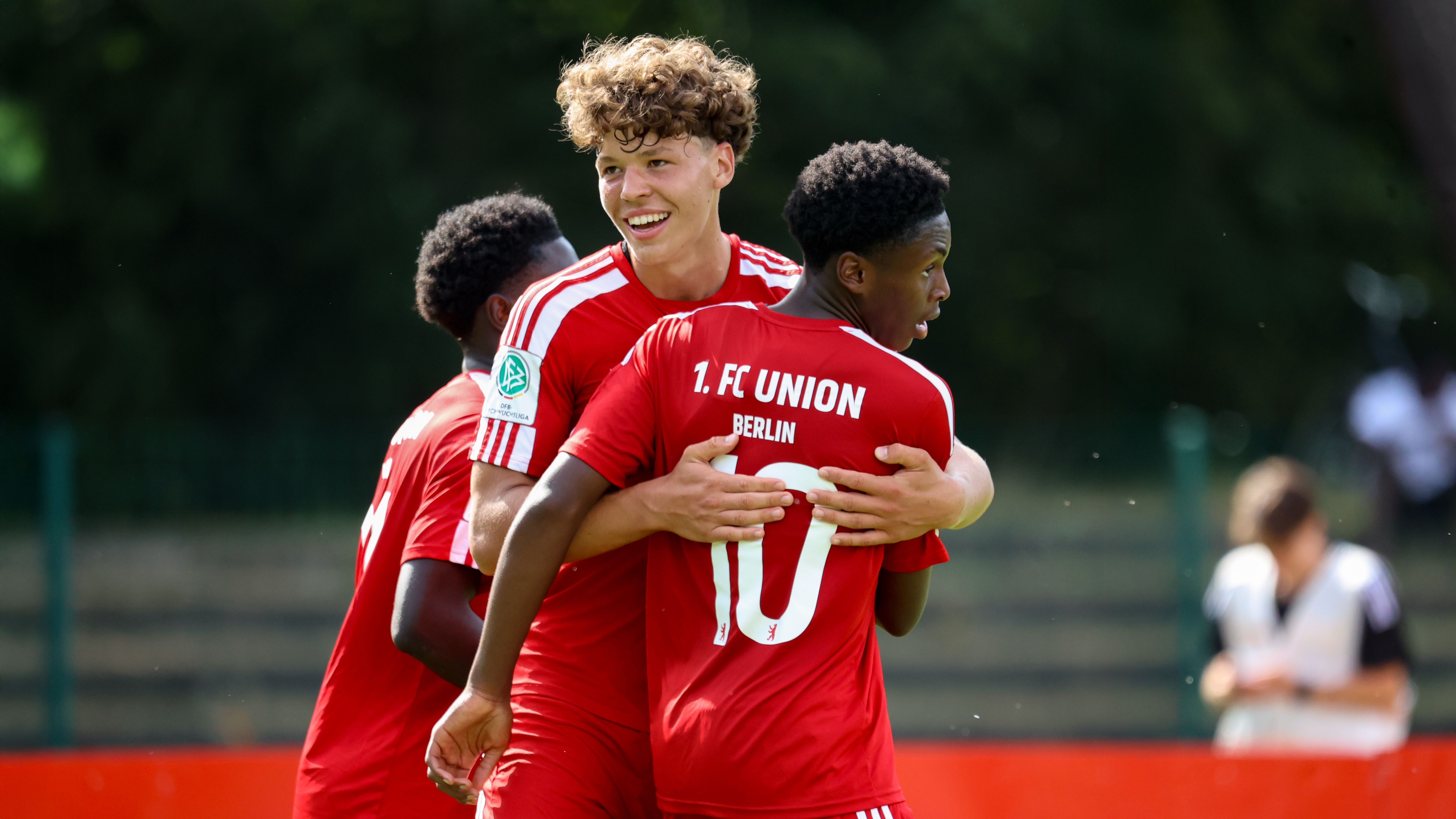 Zwei Fußballspieler in roten Trikots umarmen sich freudig, während ein weiterer Spieler im Hintergrund steht.