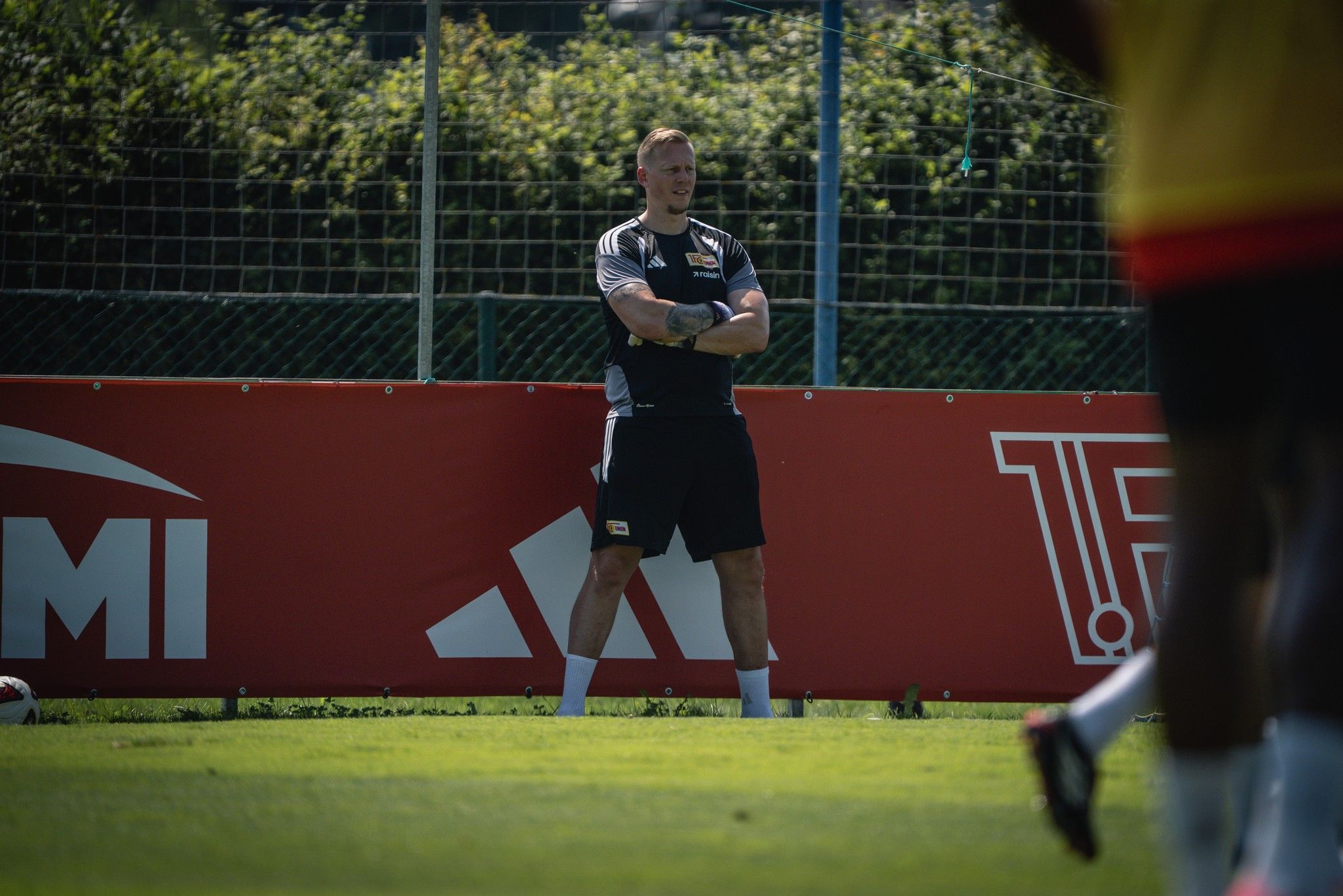 Ein Fußballtrainer steht mit verschränkten Armen auf dem Trainingsfeld neben einem Werbebanner.