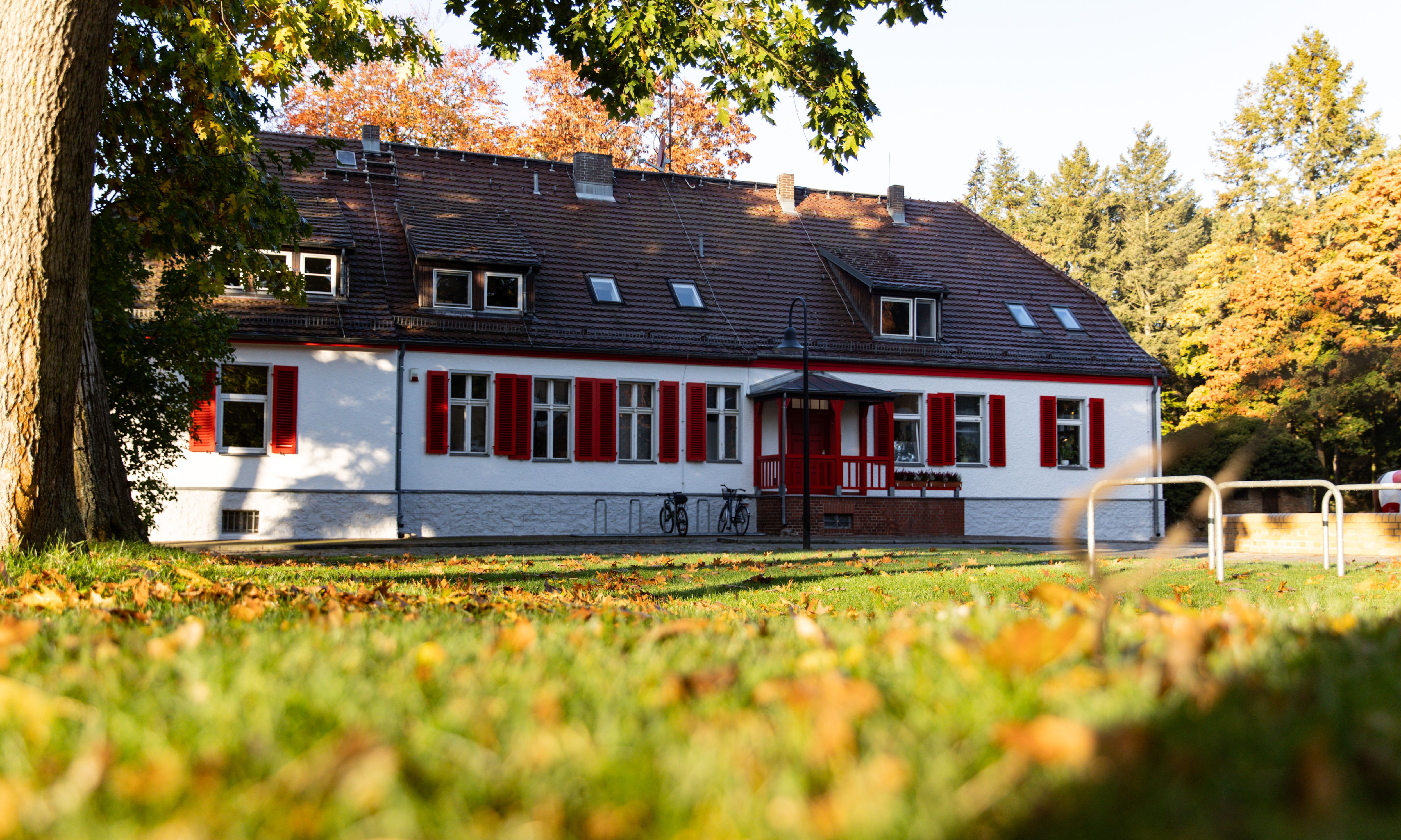 Ein zweigeschossiges, weißes Gebäude mit roten Fensterläden, umgeben von Bäumen und herbstlichen Blättern.