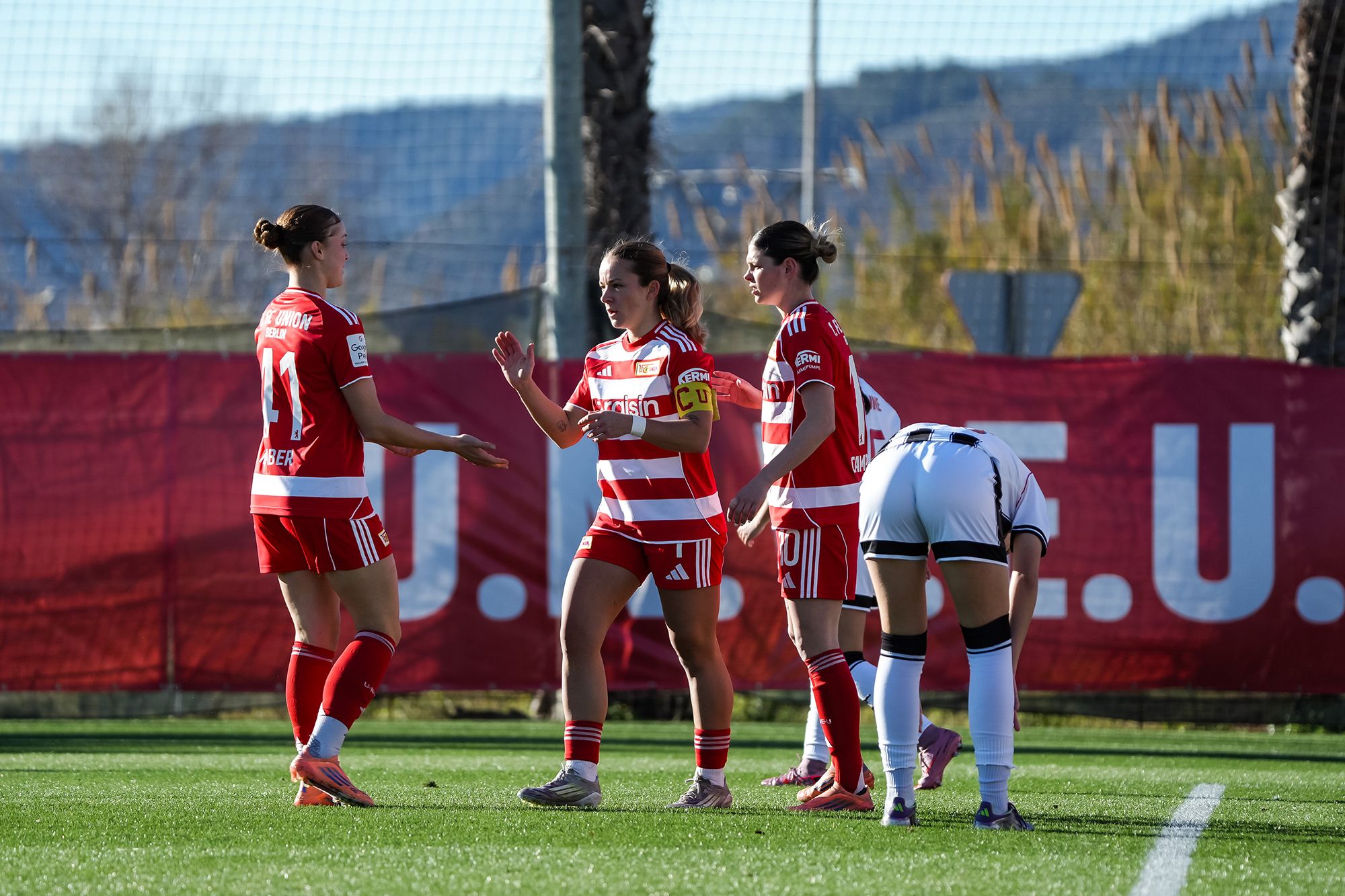 Drei Fußballspielerinnen in rot-weiß gestreiften Trikots feiern einen Erfolg auf dem Spielfeld. Im Hintergrund ist ein Tor sichtbar.